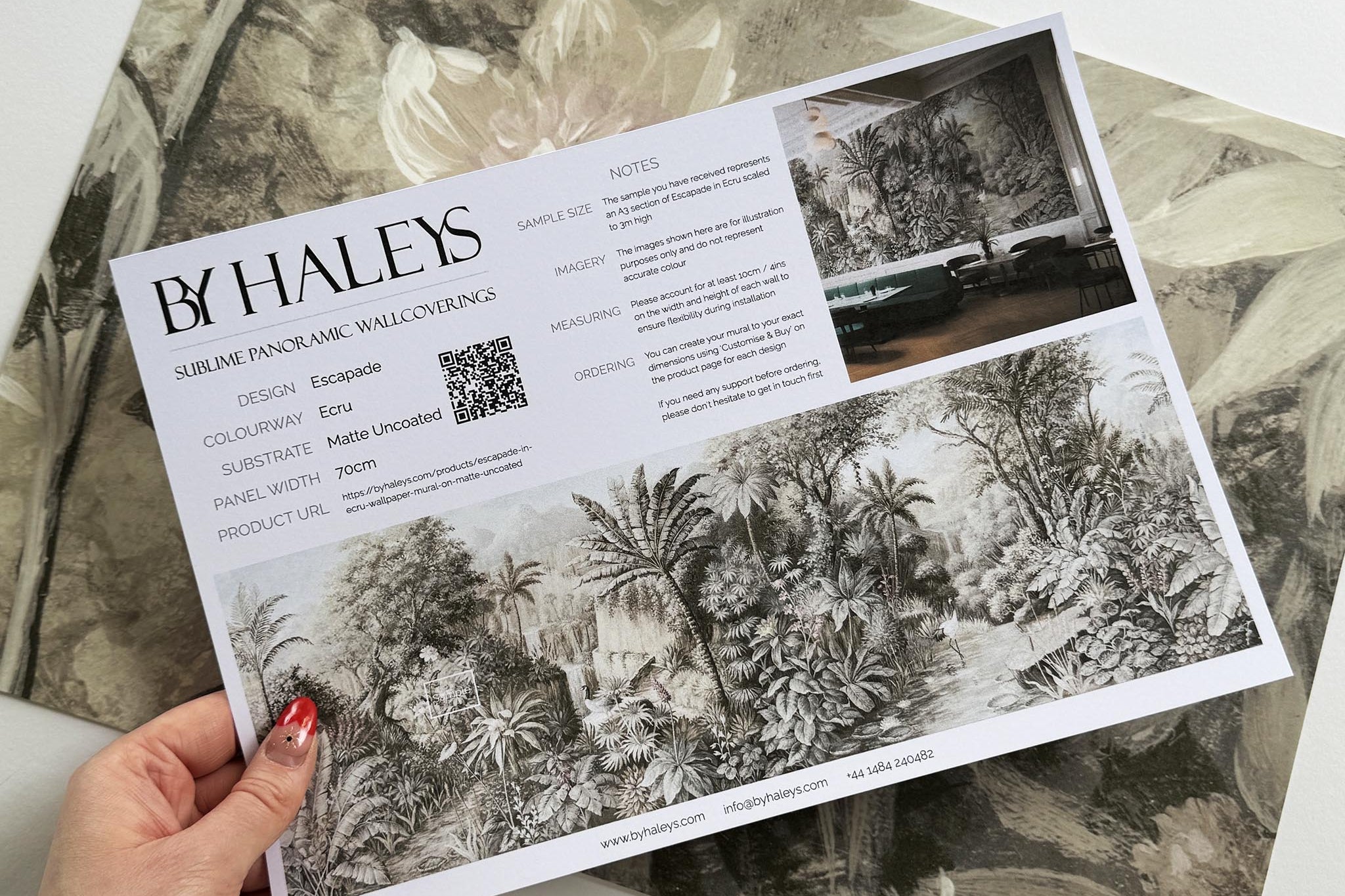 A hand showing information for a matte wallpaper mural sample which features a jungle vista in sepia and blush.