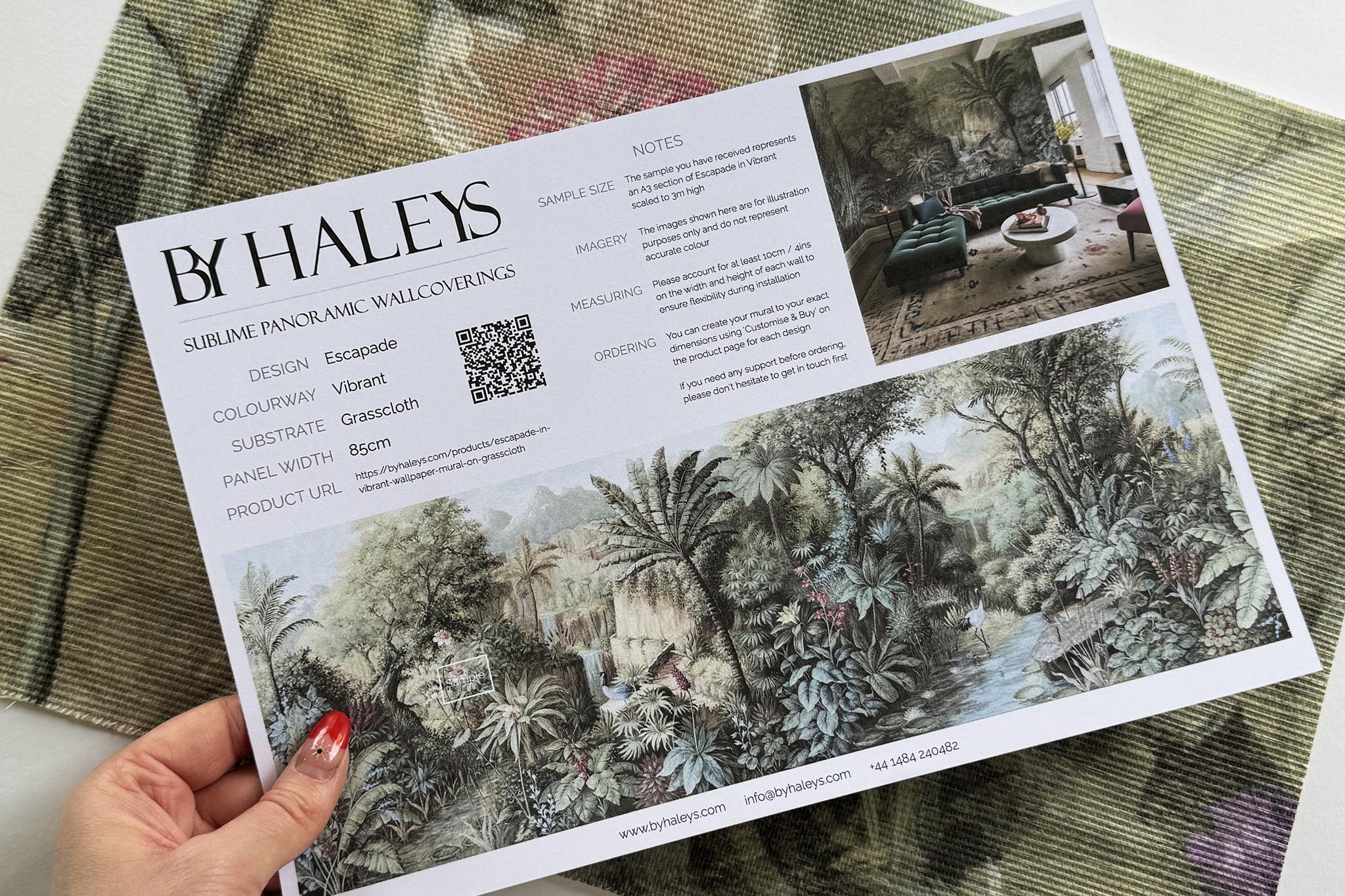 A hand showing information for a grasscloth wallpaper mural sample which features a tropical paradise in cool greens and blushes.