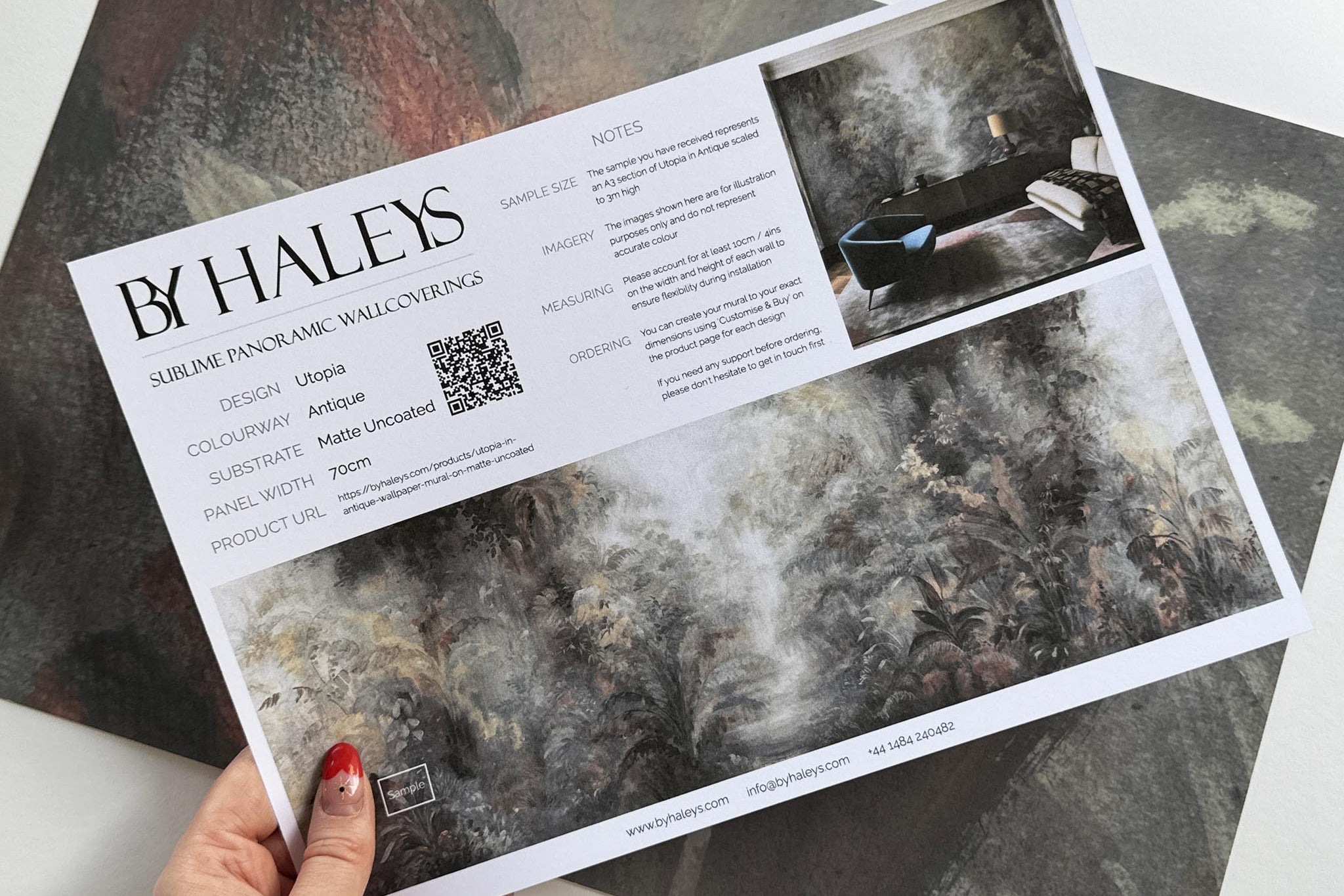A hand showing information for a matte wallpaper mural sample which features a rainforest in lavender and rust.