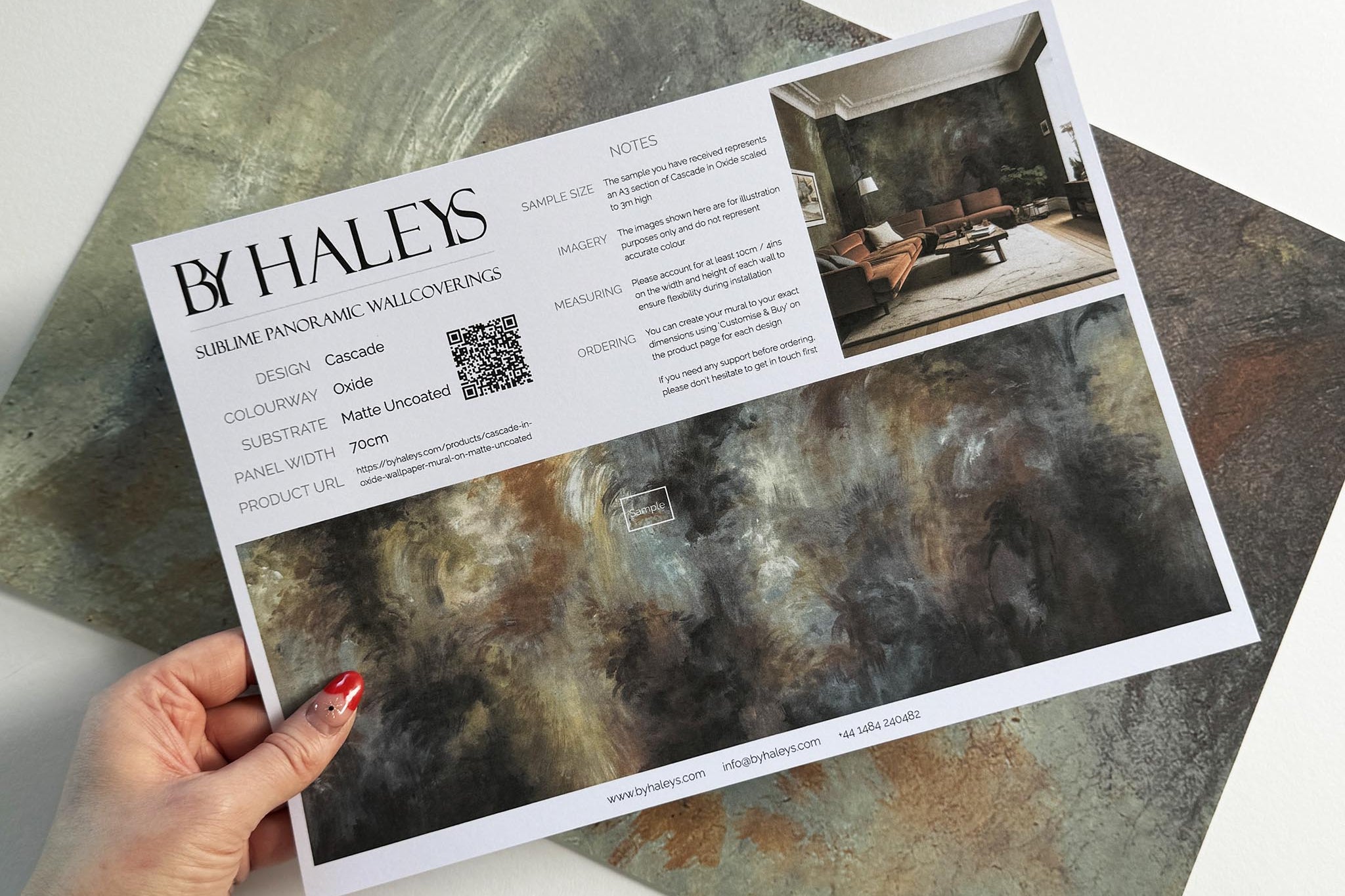 A hand showing information for a matte wallpaper mural sample which features an abstract haze in charcoal and ash.