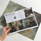 A hand showing information for a matte wallpaper mural sample which features an abstract haze in charcoal and ash.
