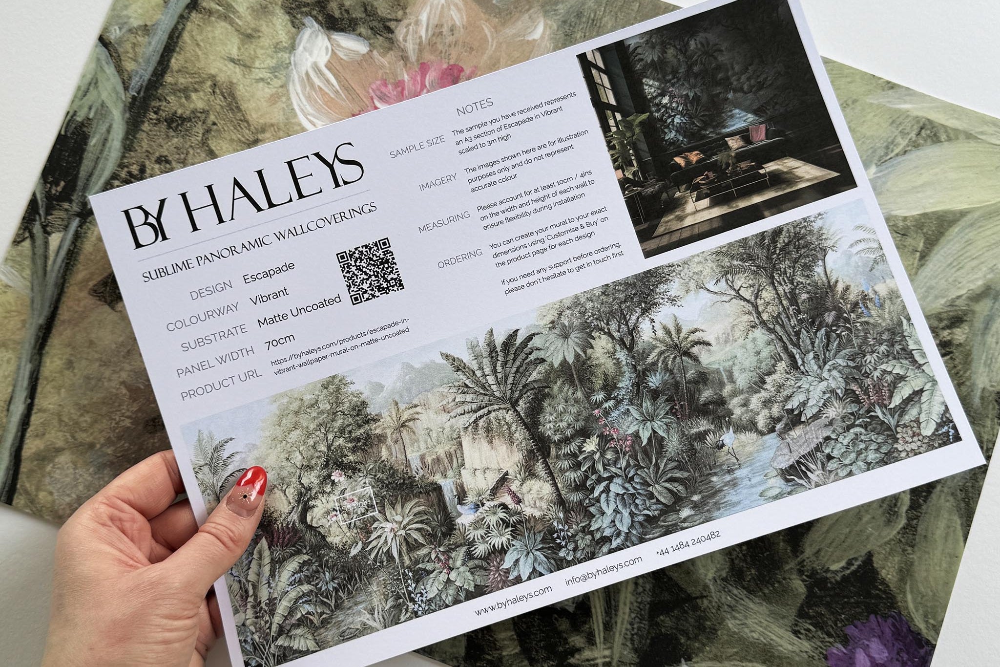 A hand showing information for a matte wallpaper mural sample which features a tropical paradise in cool greens and blushes.