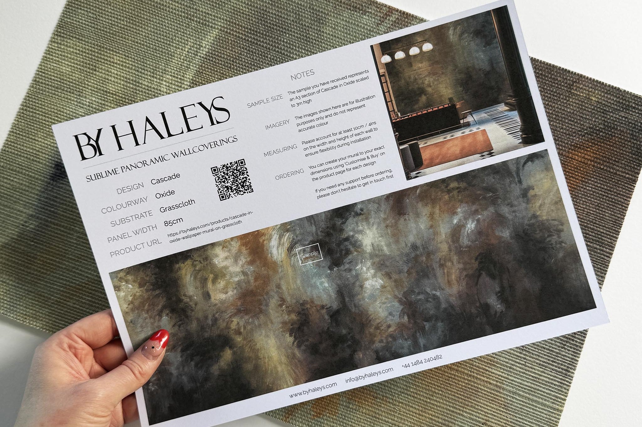 A hand showing information for a grasscloth wallpaper mural sample which features an abstract haze in charcoal and ash.