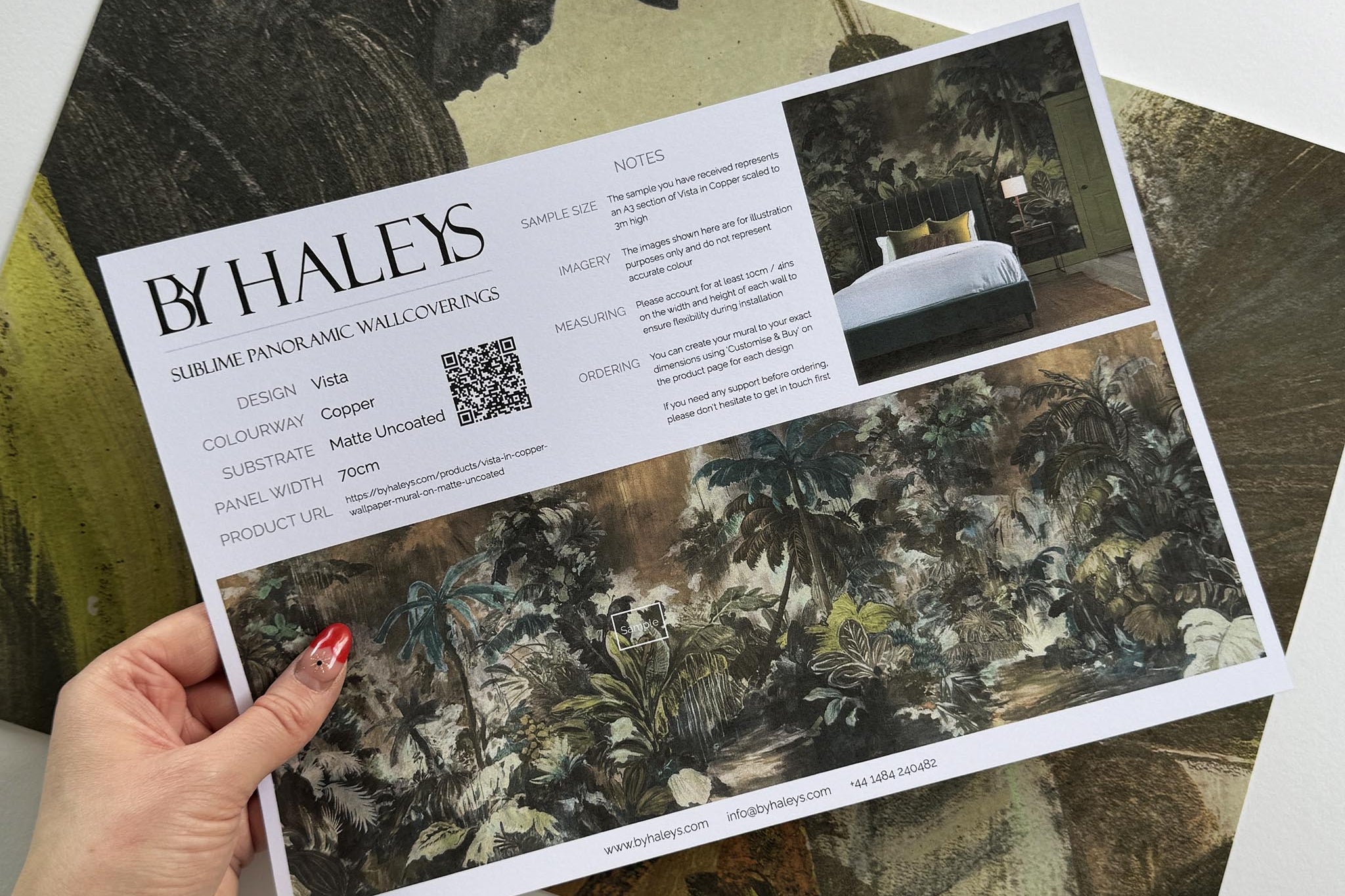 A hand showing information for a matte wallpaper mural sample which features a moody jungle in teal and ochre.