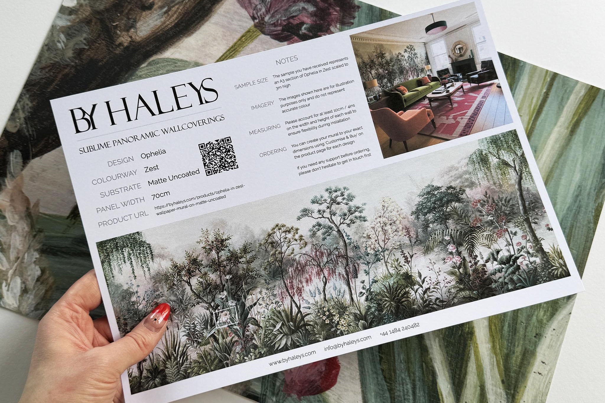 A hand showing information for a matte wallpaper mural sample which features a lakeside garden scene in mint and pink.