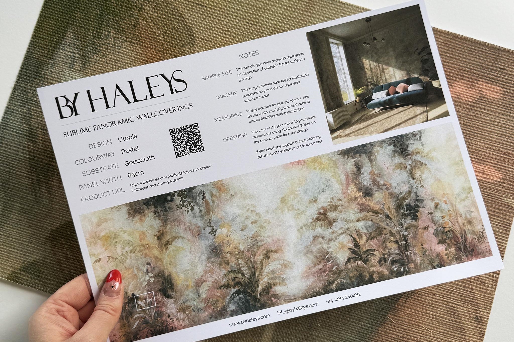A hand showing information for a grasscloth wallpaper mural sample which features a rainforest in blush and sky blue.