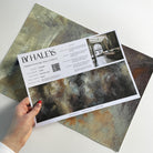 A hand showing information for a vinyl wallpaper mural sample which features an abstract haze in charcoal and ash.