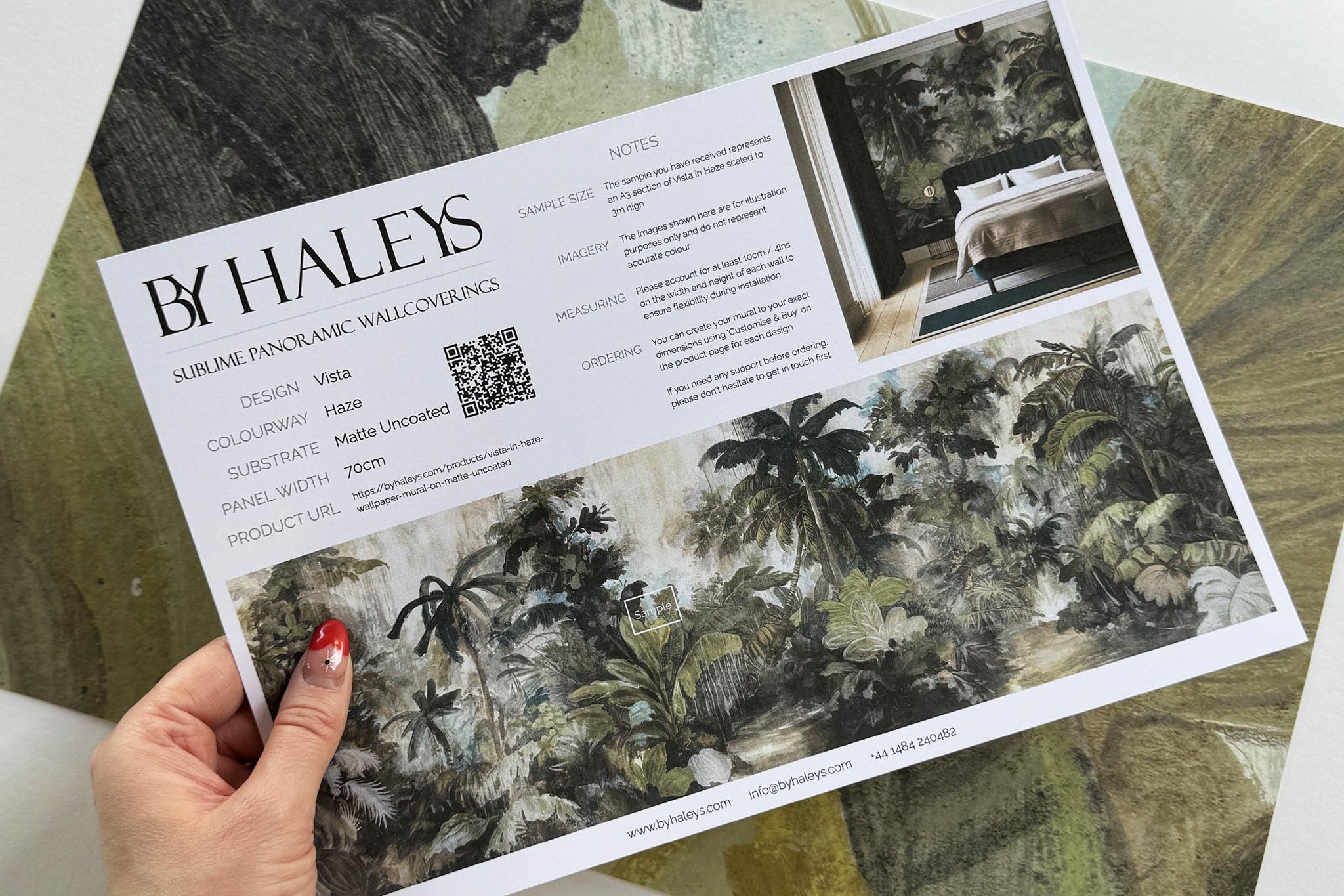 A hand showing information for a matte wallpaper mural sample which features a tropical scene in sage and parchment.