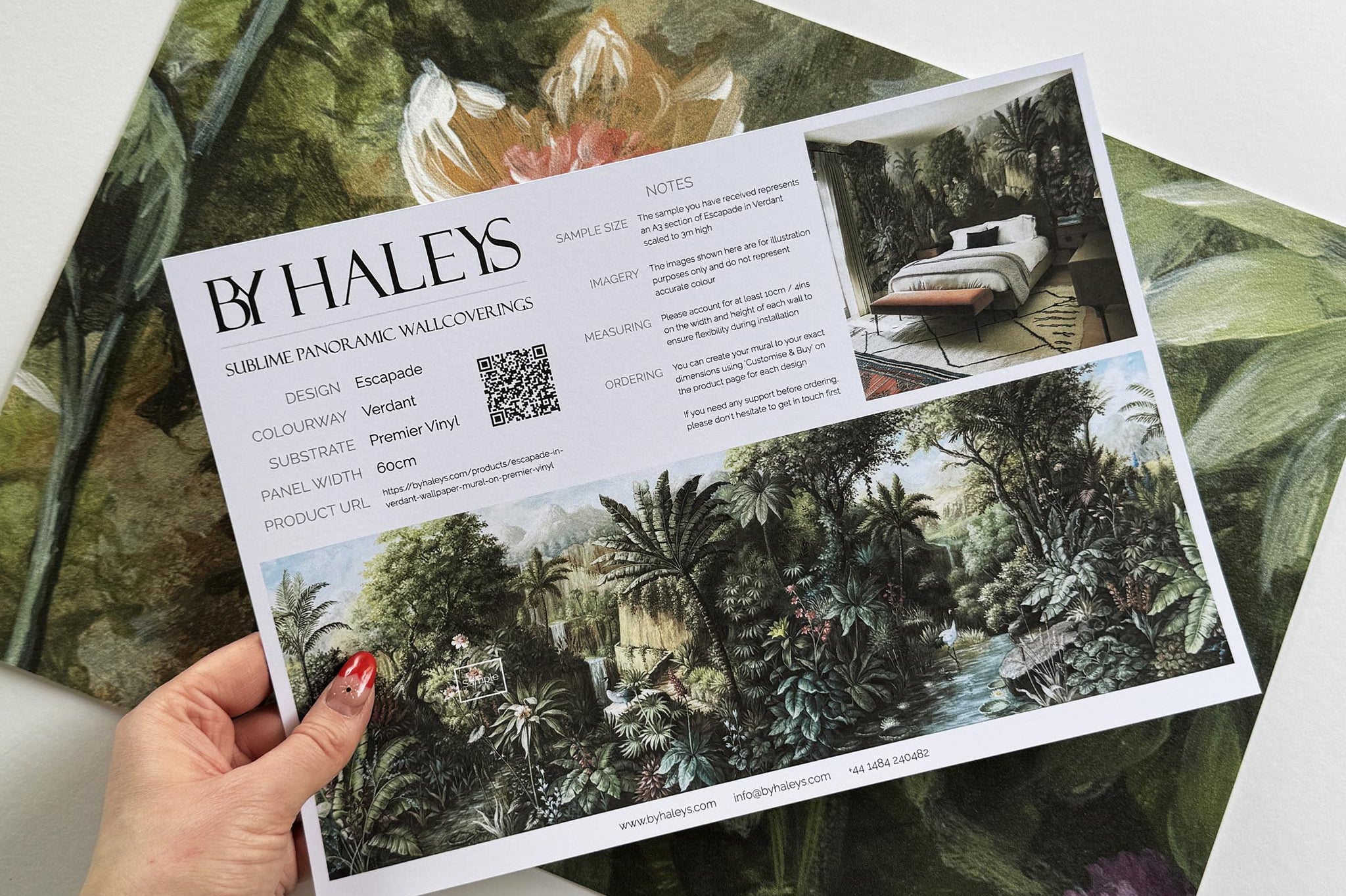 A hand showing information for a vinyl wallpaper mural sample which features a tropical scene in green and pink.