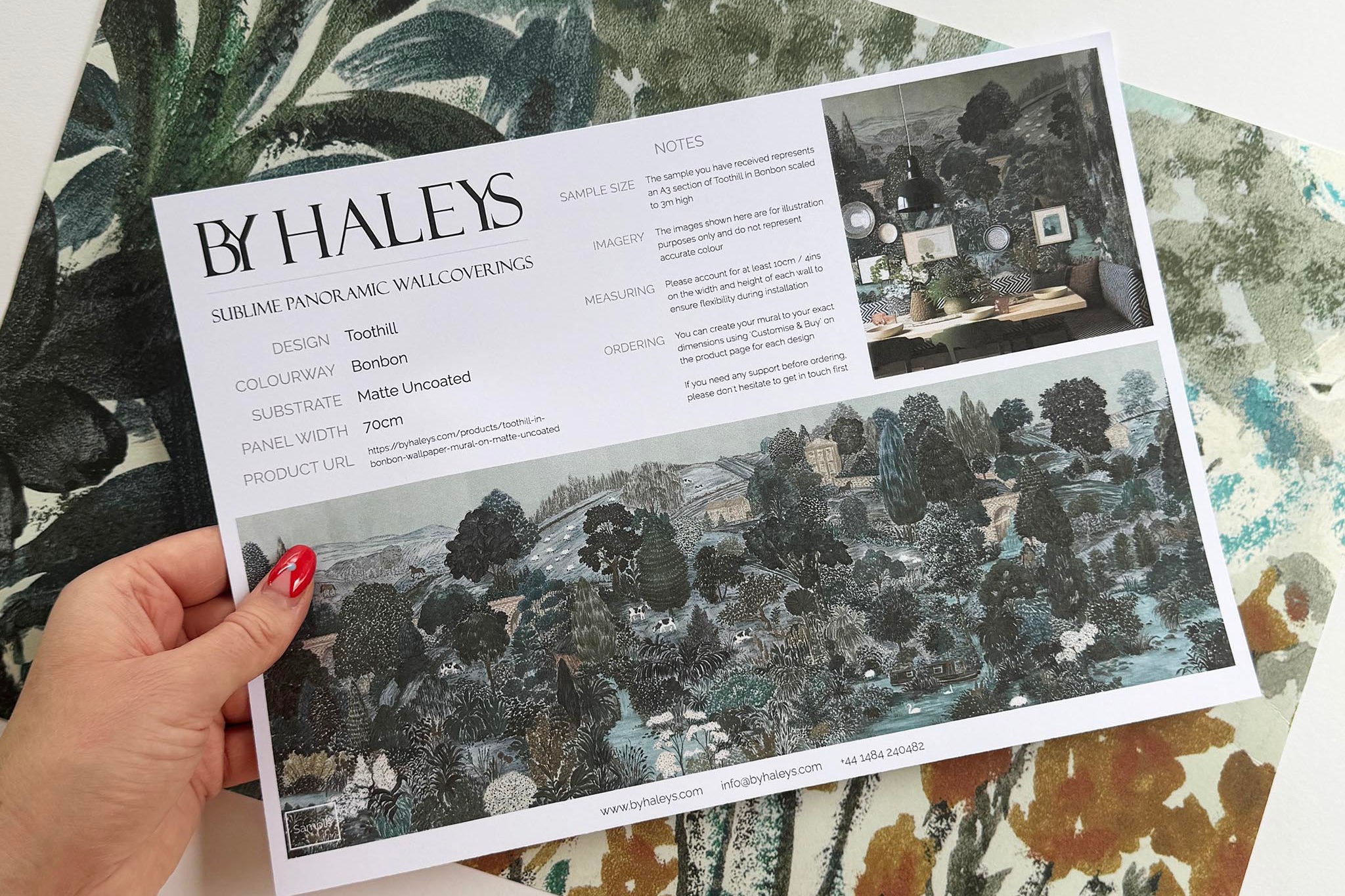A hand showing information for a matte wallpaper mural sample which features a countryside in teal, sea green and slate