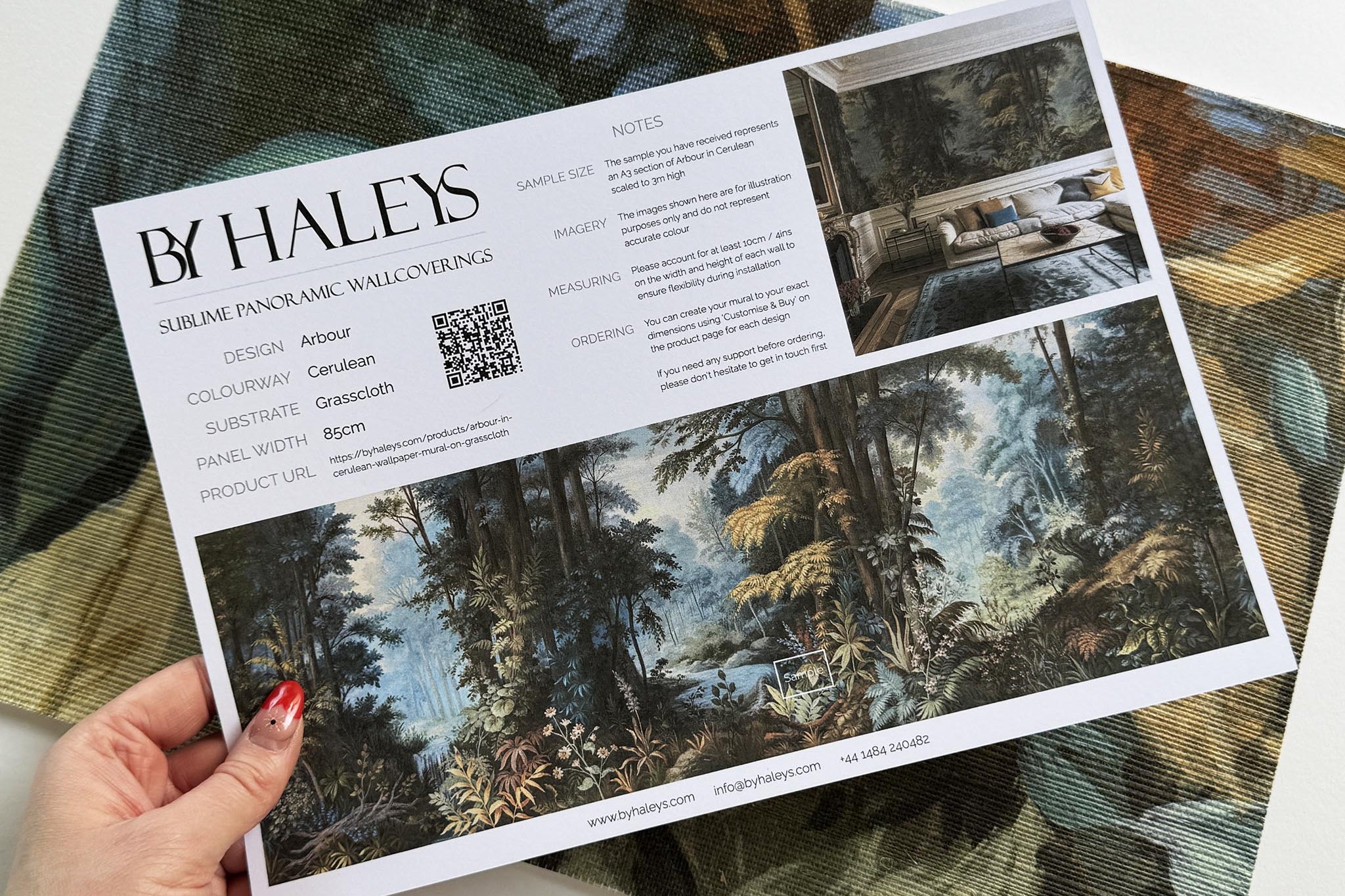 A hand showing information for a grasscloth wallpaper mural sample which features an enchanted forest in blue and rust.