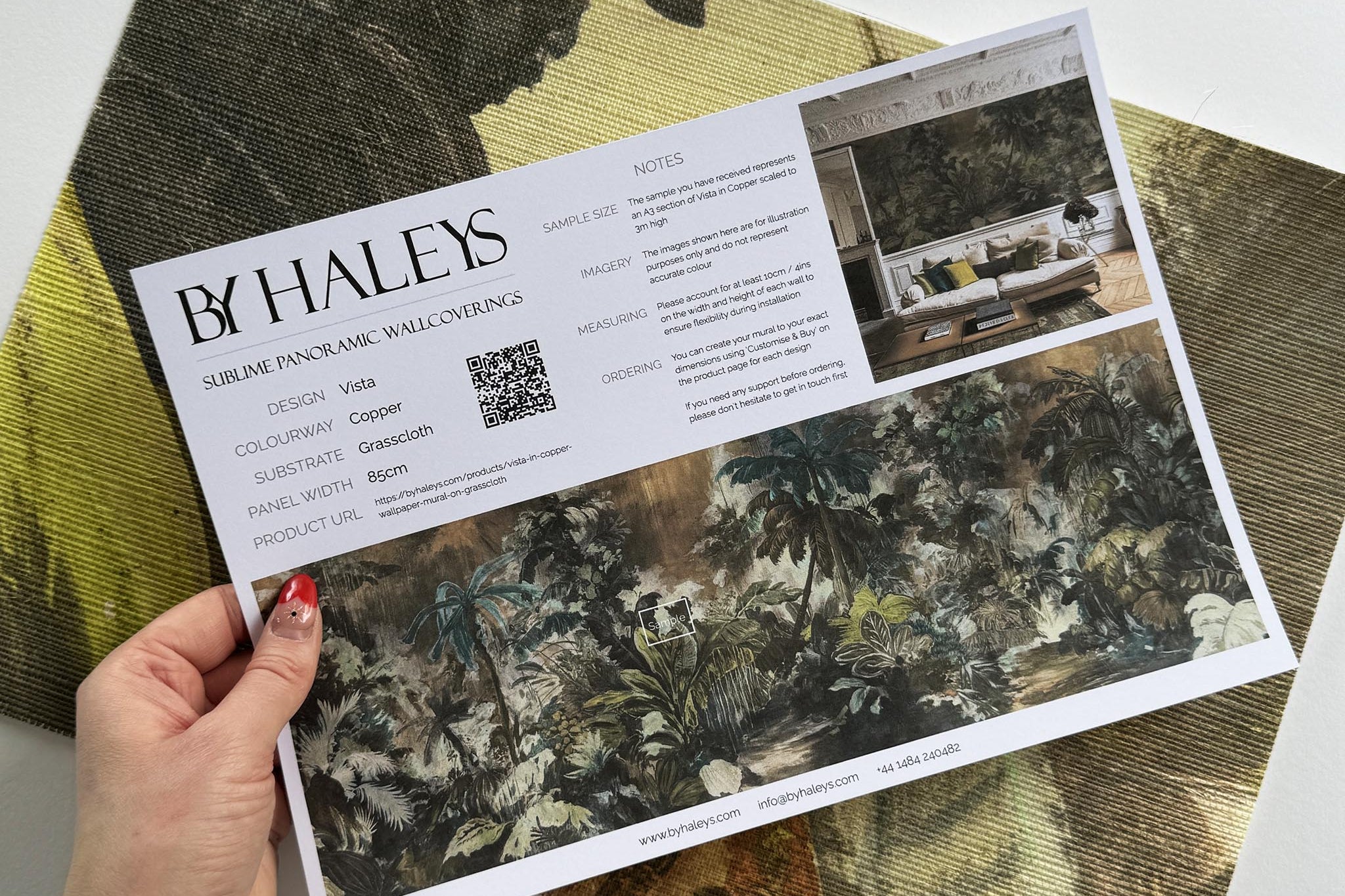 A hand showing information for a grasscloth wallpaper mural sample which features a moody jungle in teal and ochre.