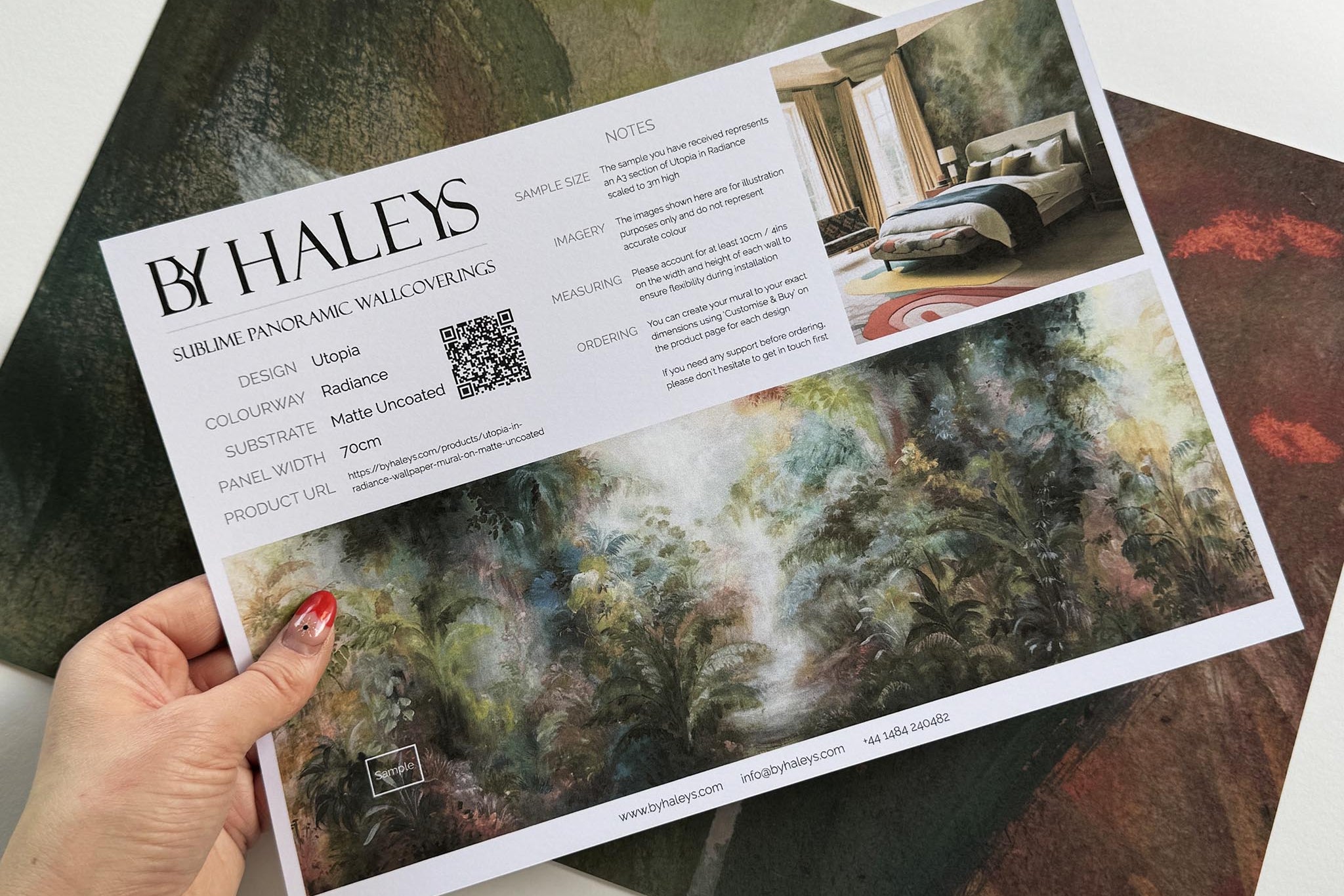 A hand showing information for a matte wallpaper mural sample which features a rainforest in green, coral, and gold.
