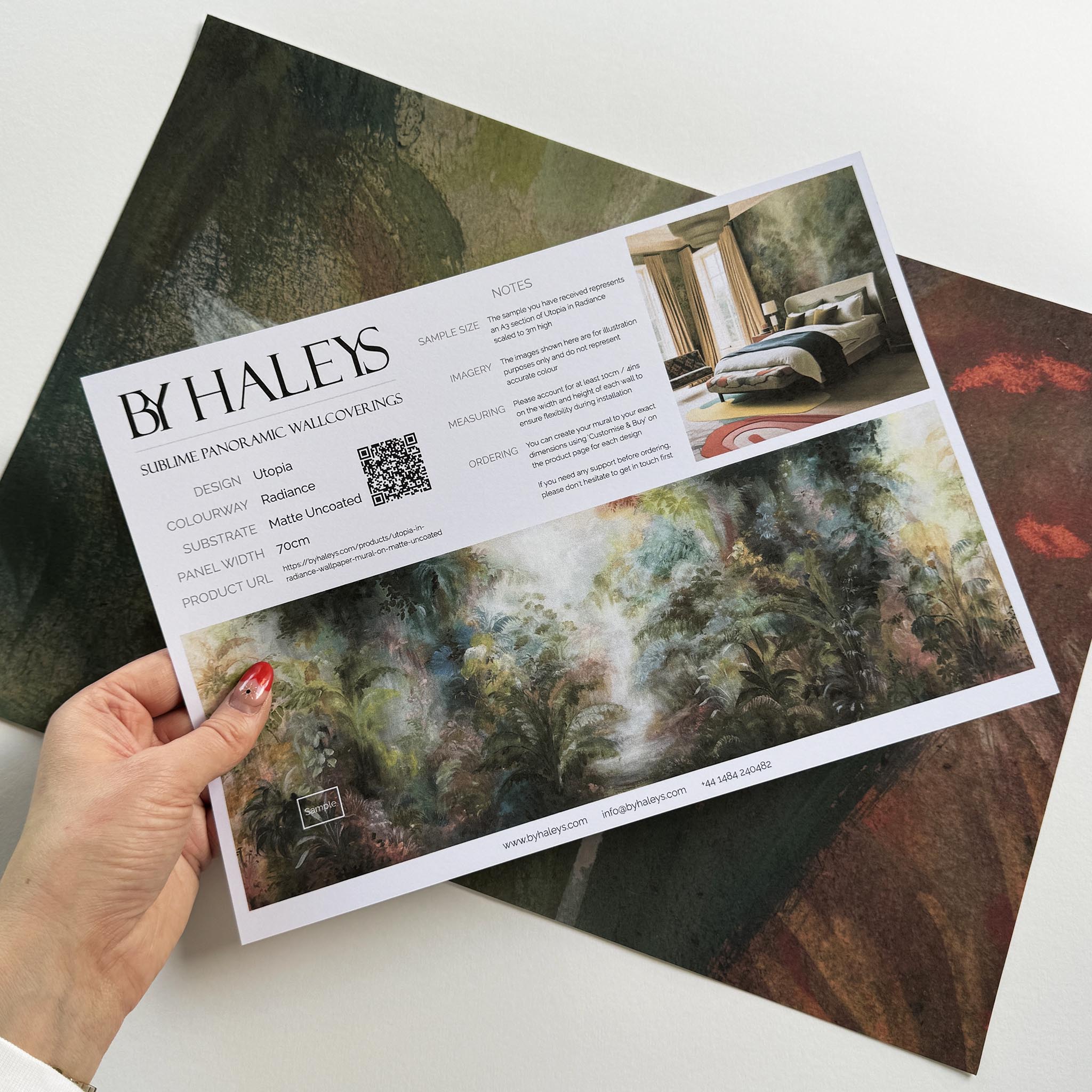 A hand showing information for a matte wallpaper mural sample which features a rainforest in green, coral, and gold.