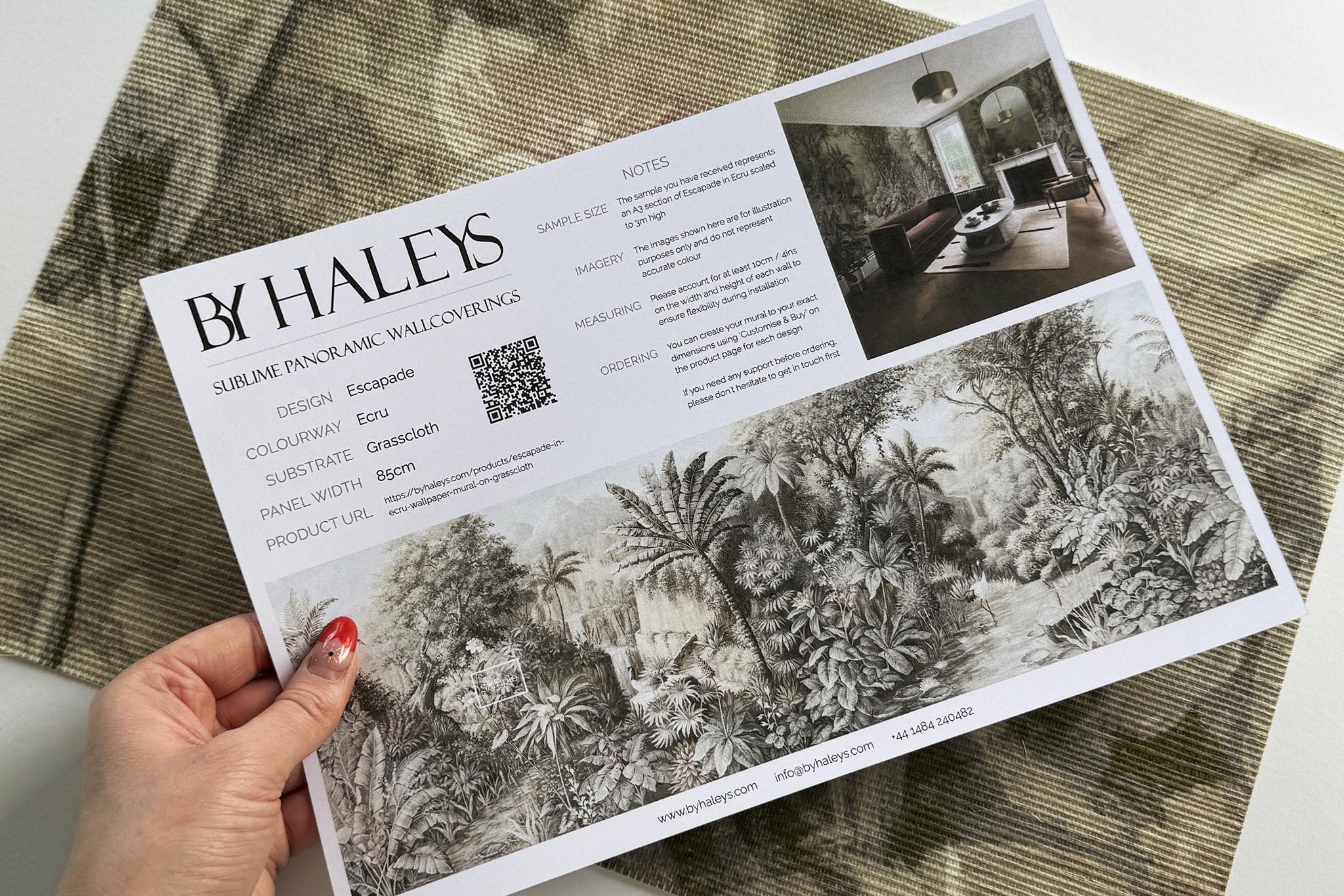 A hand showing information for a grasscloth wallpaper mural sample which features a jungle vista in sepia and blush.