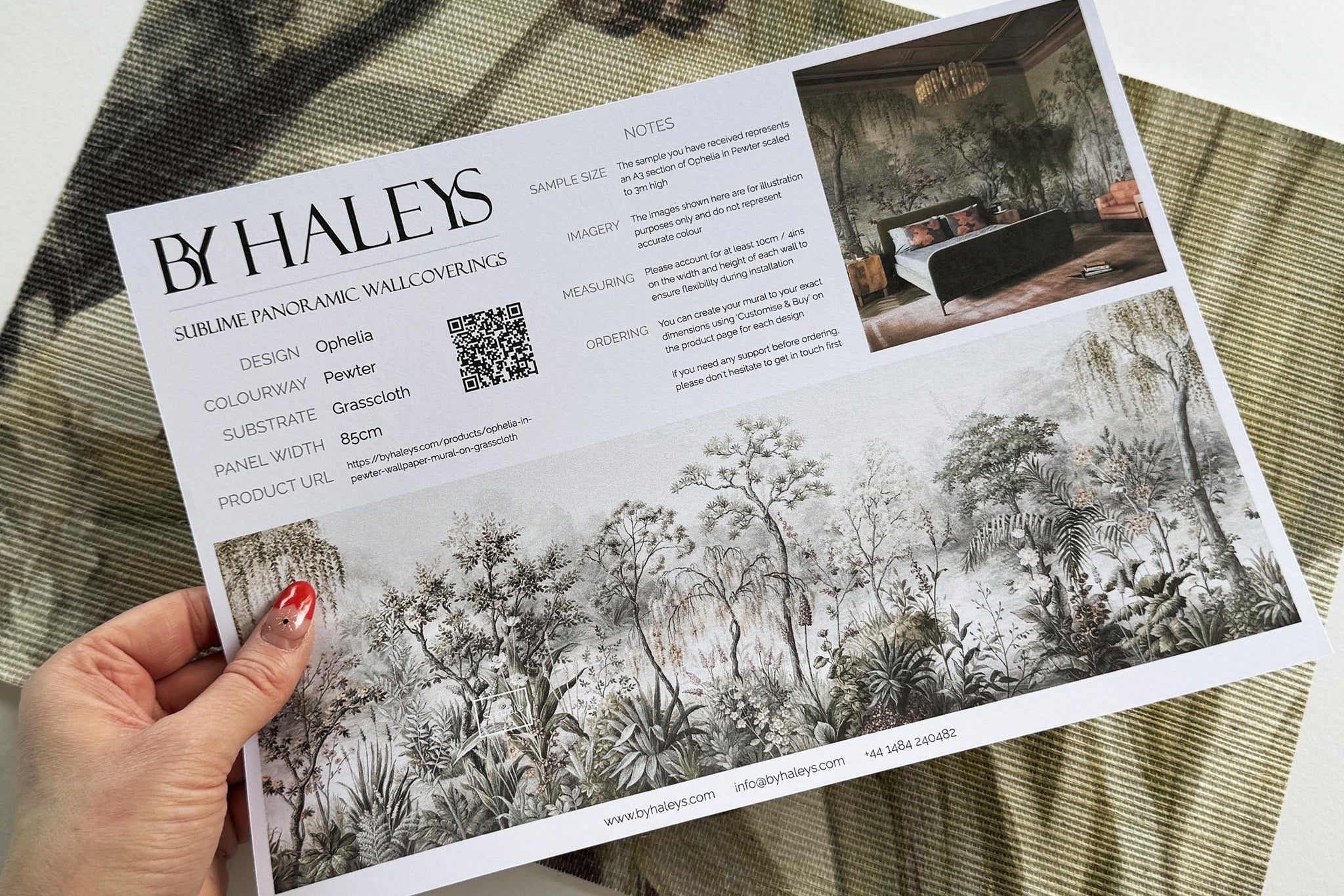 A hand showing information for a grasscloth wallpaper mural sample which features a lakeside scene in grey and peach.