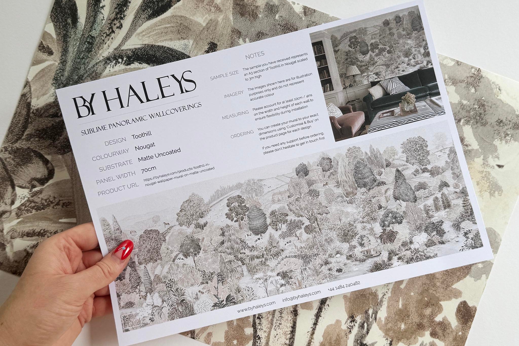 A hand showing information for a matte wallpaper mural sample which features a countryside in almond, dusty rose, and mist