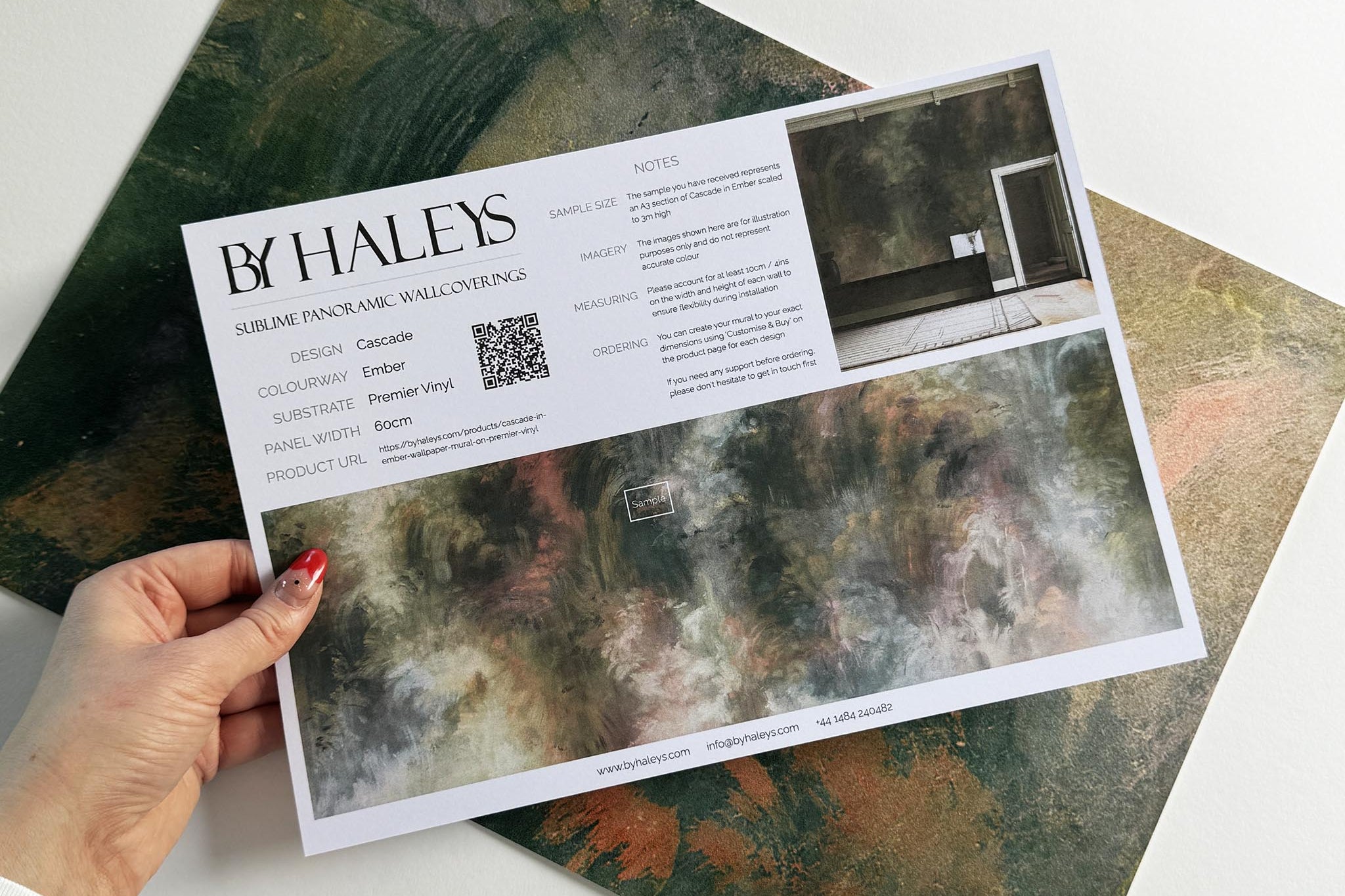 A hand showing information for a vinyl wallpaper mural sample which features an abstract haze in green and blush.