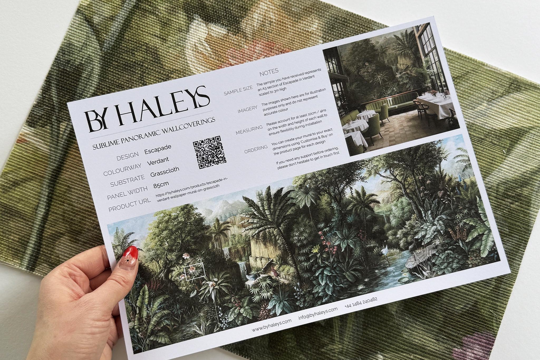 A hand showing information for a grasscloth wallpaper mural sample which features a tropical scene in green and pink.