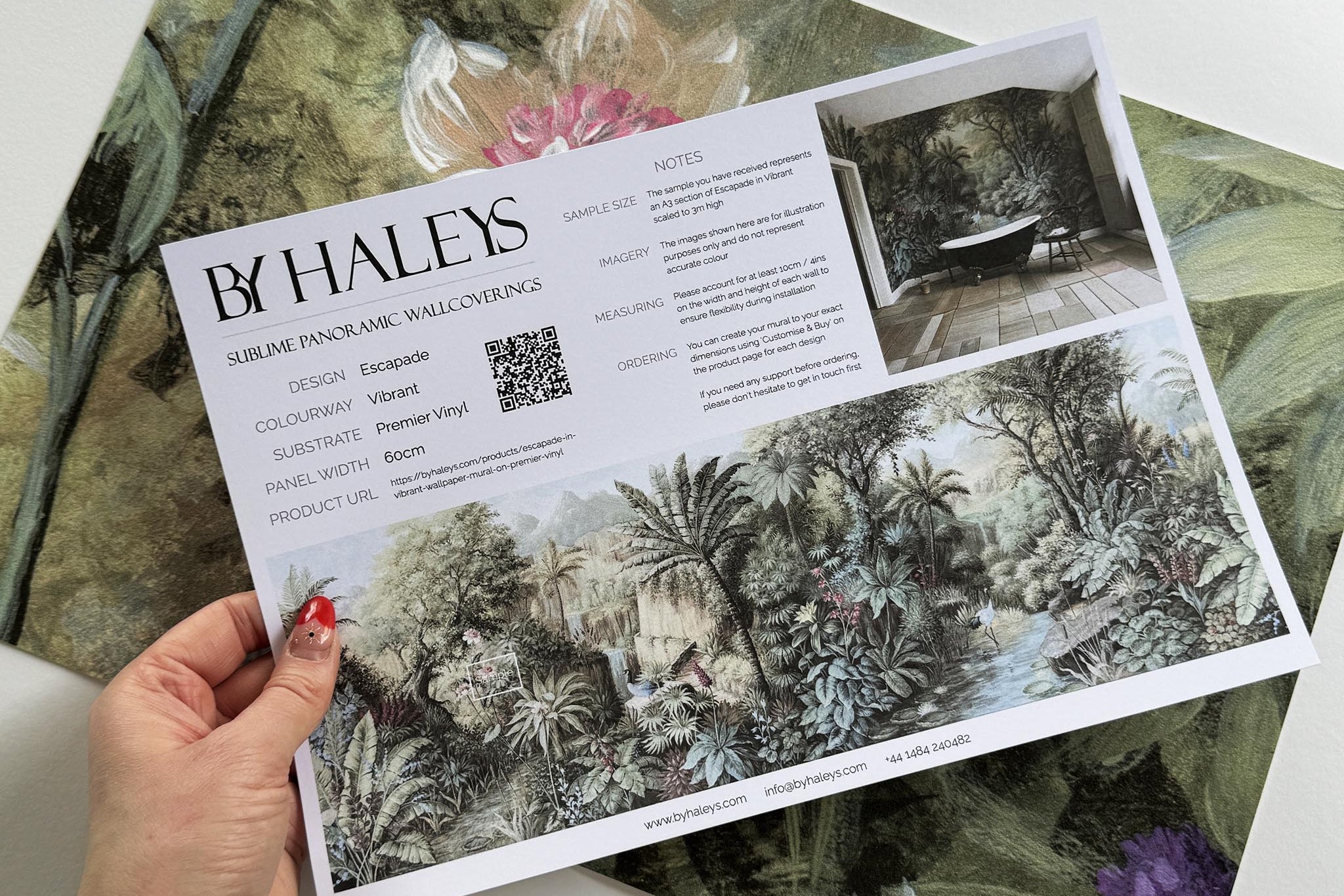 A hand showing information for a vinyl wallpaper mural sample which features a tropical paradise in cool greens and blushes.