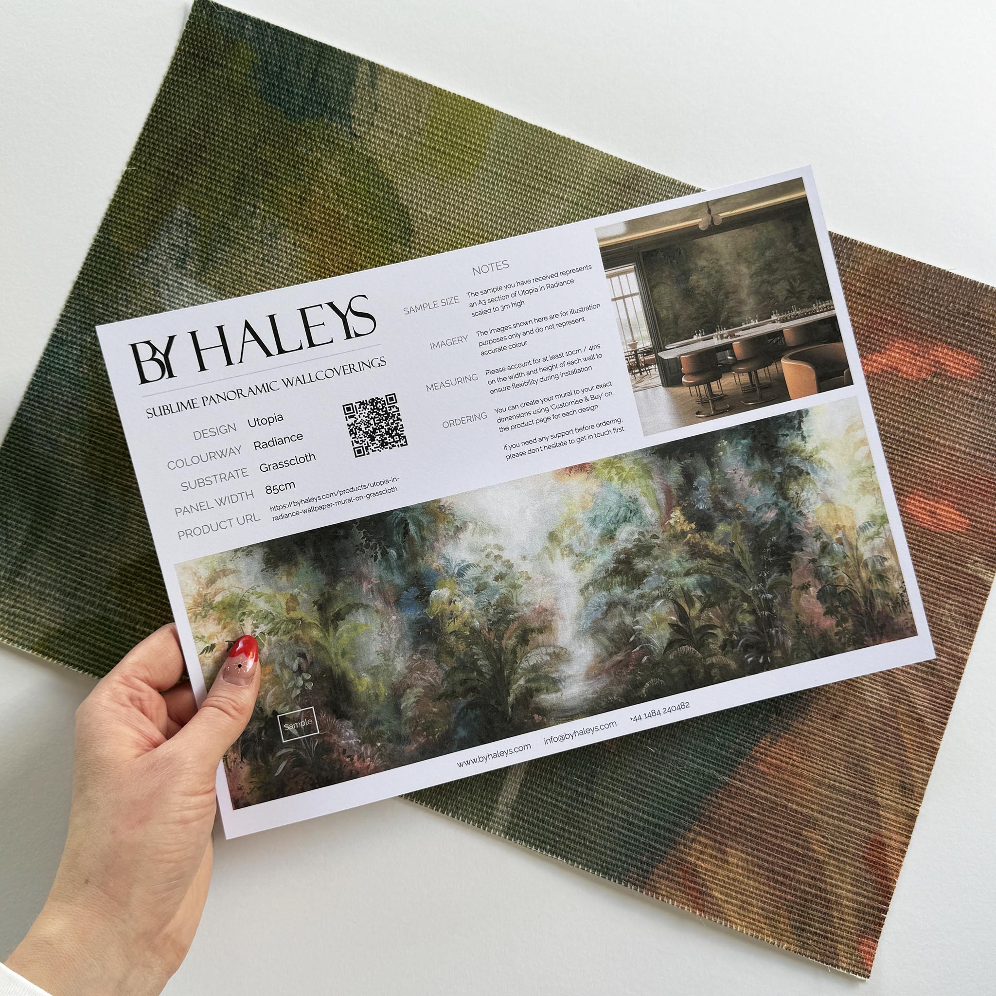 A hand showing information for a grasscloth wallpaper mural sample which features a rainforest in green, coral, and gold.