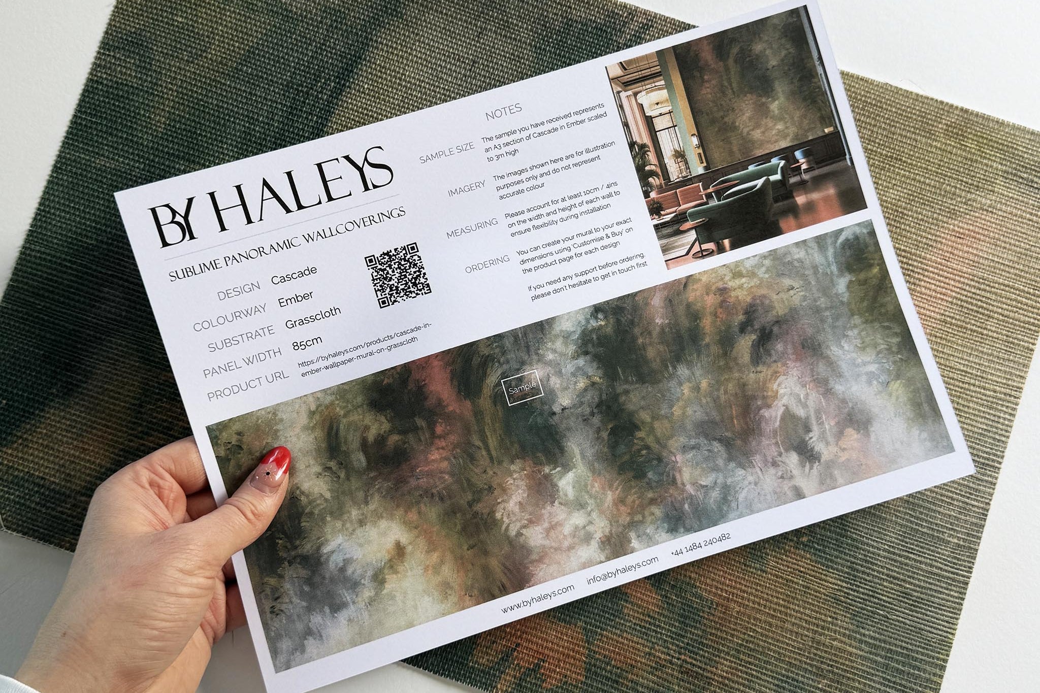 A hand showing information for a grasscloth wallpaper mural sample which features an abstract haze in green and blush.