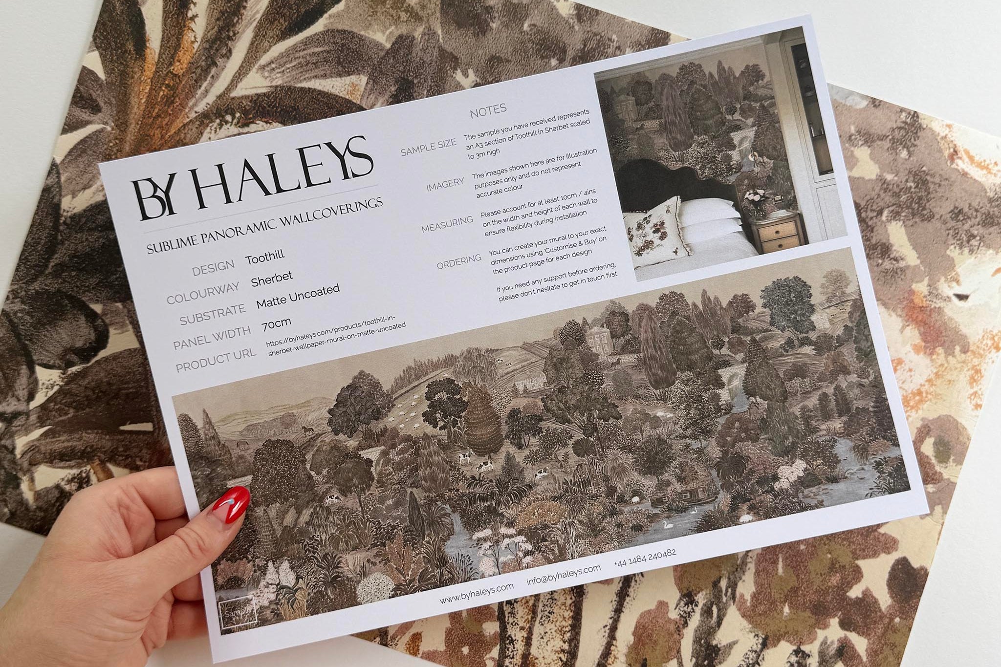 A hand showing information for a matte wallpaper mural sample which features a countryside in clay and soft plum tones