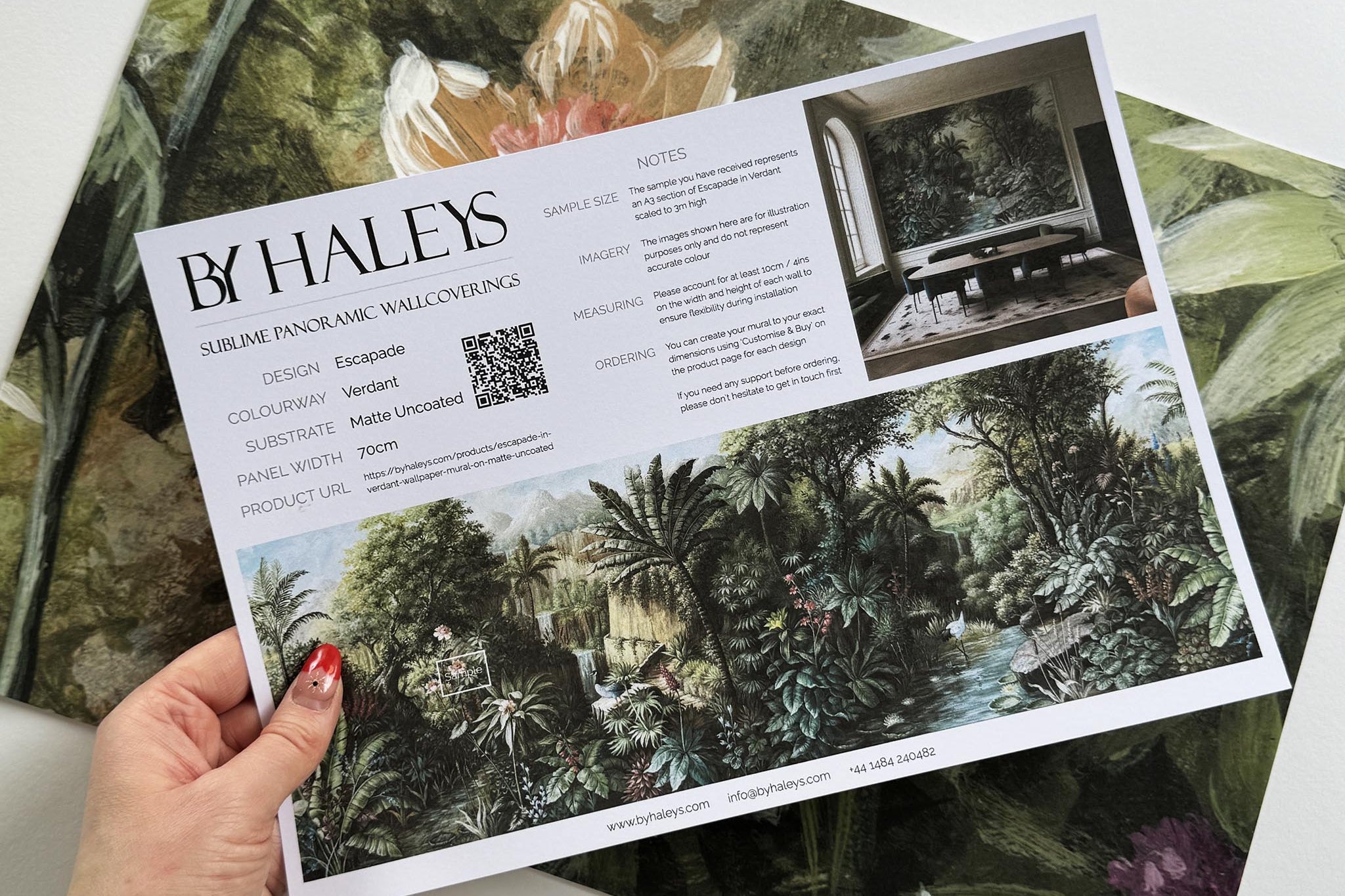 A hand showing information for a matte wallpaper mural sample which features a tropical scene in green and pink.