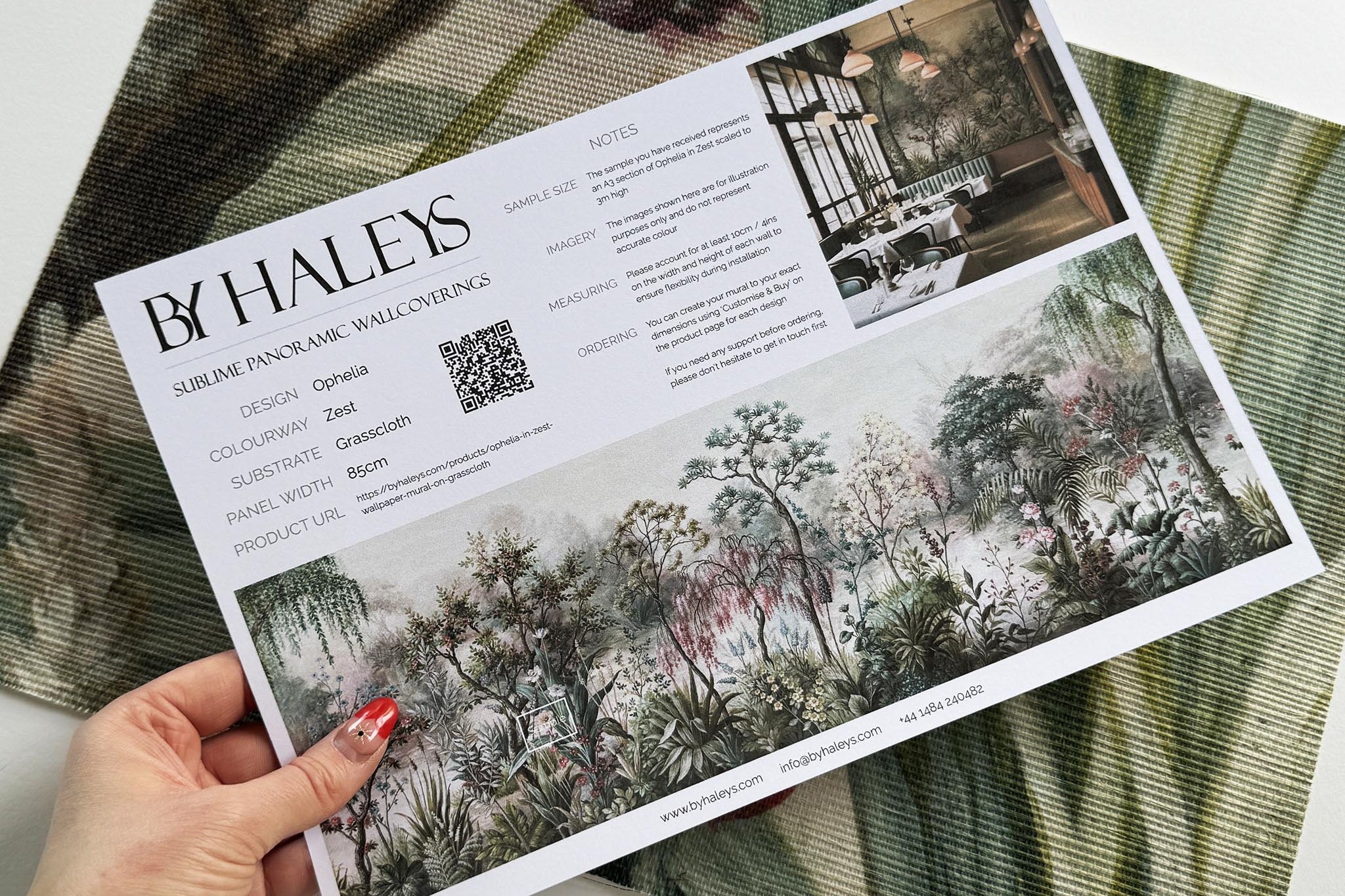 A hand showing information for a grasscloth wallpaper mural sample which features a lakeside garden scene in mint and pink.