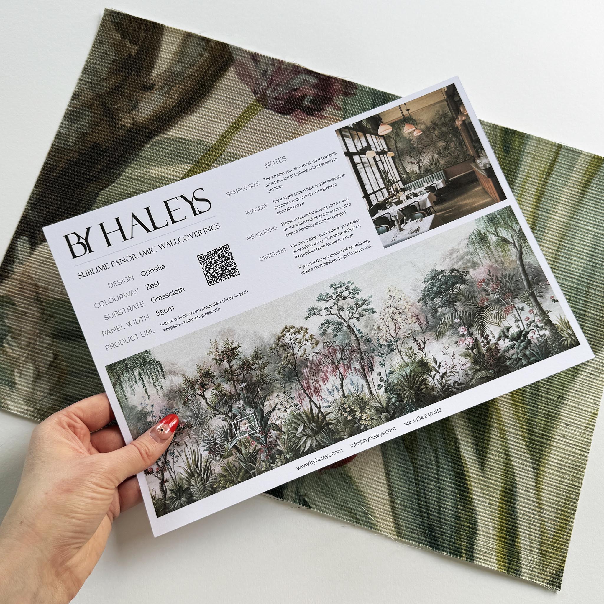 A hand showing information for a grasscloth wallpaper mural sample which features a lakeside garden scene in mint and pink.
