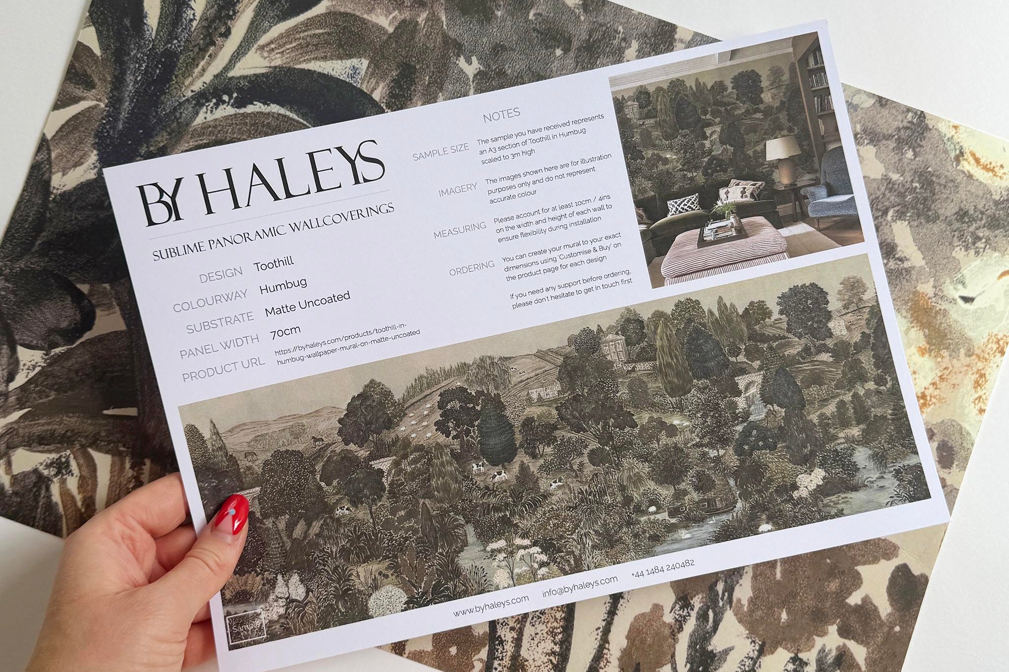 A hand showing information for a matte wallpaper mural sample which features a countryside in deep olive, slate and ash