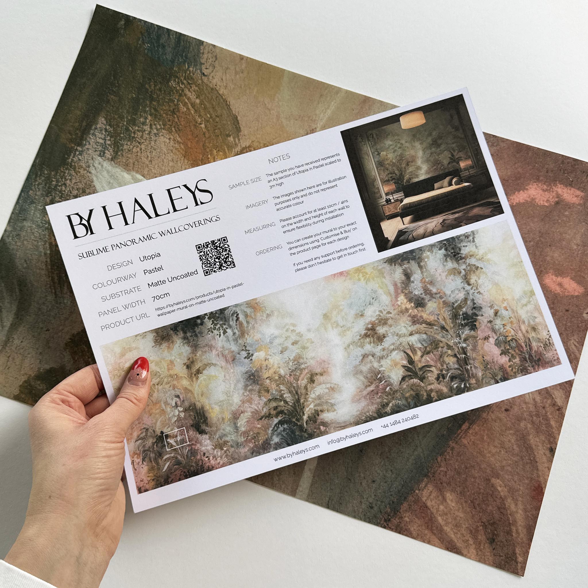 A hand showing information for a matte wallpaper mural sample which features a rainforest in blush and sky blue.