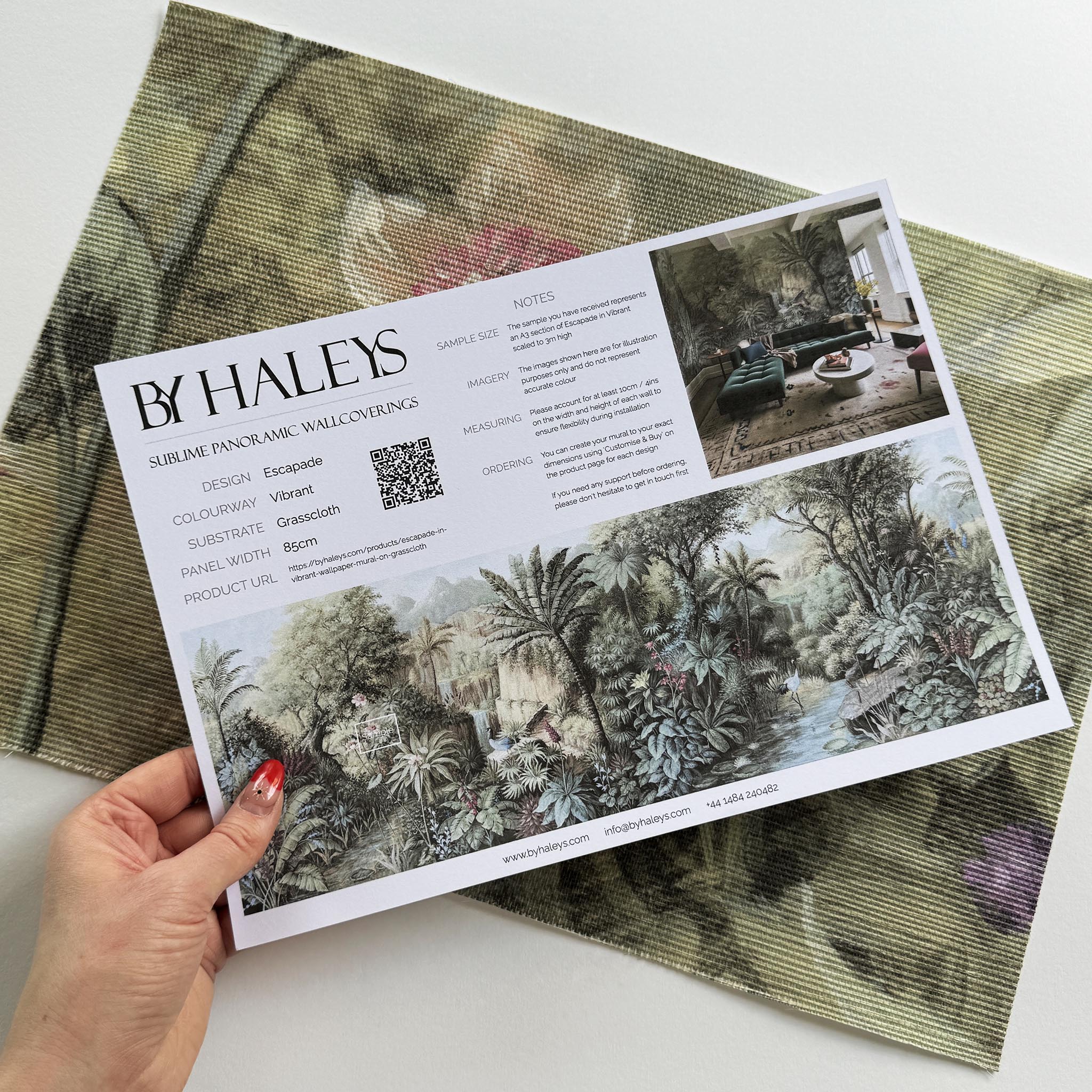 A hand showing information for a grasscloth wallpaper mural sample which features a tropical paradise in cool greens and blushes.