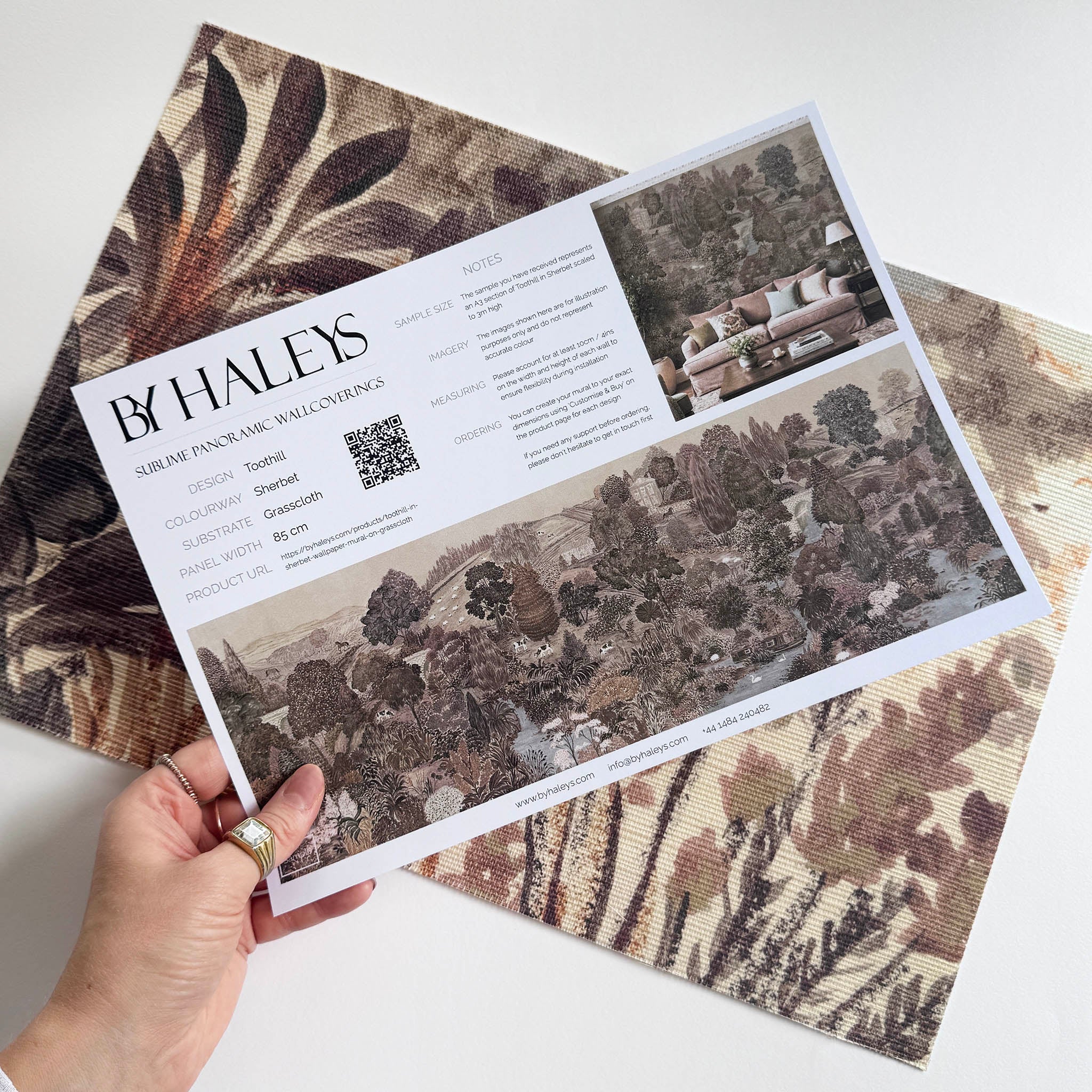 A hand showing information for a grasscloth wallpaper mural sample which features a countryside in teal, sea green and slate 