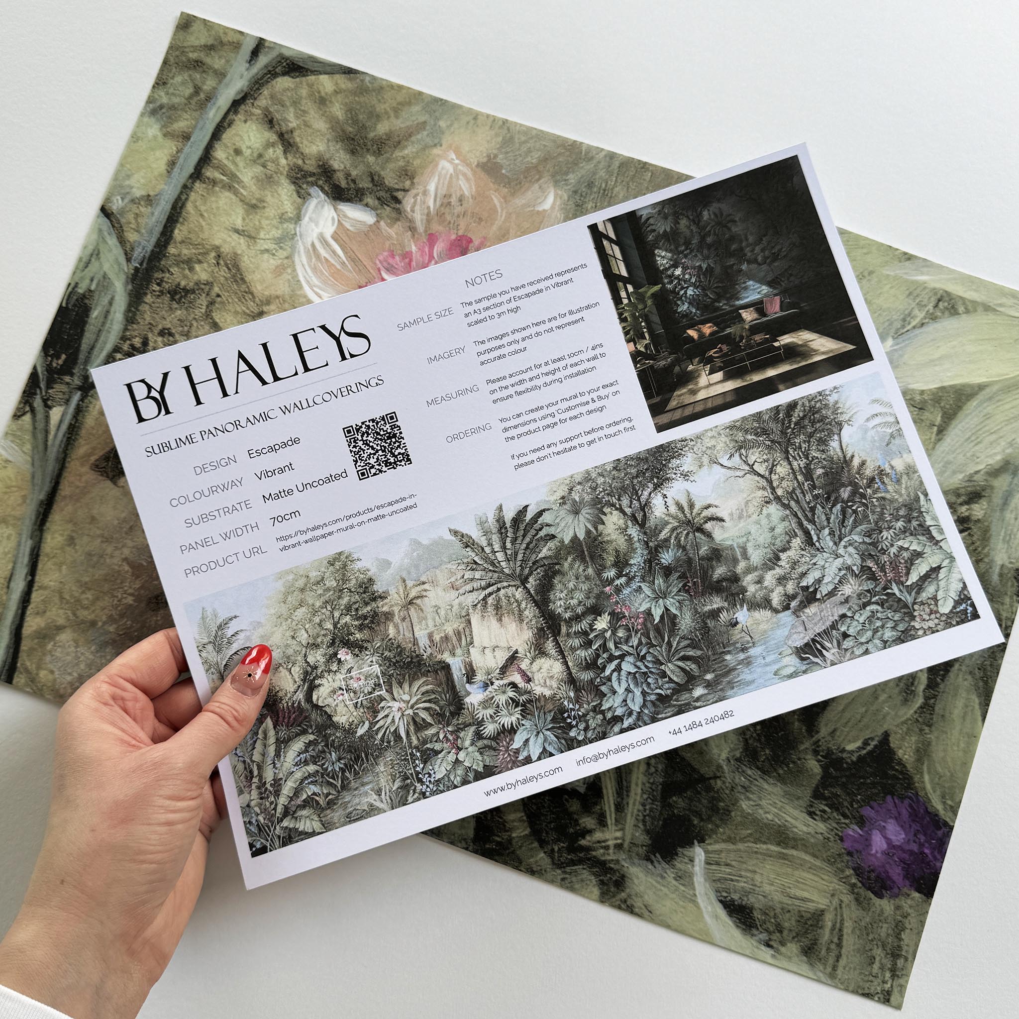 A hand showing information for a matte wallpaper mural sample which features a tropical paradise in cool greens and blushes.