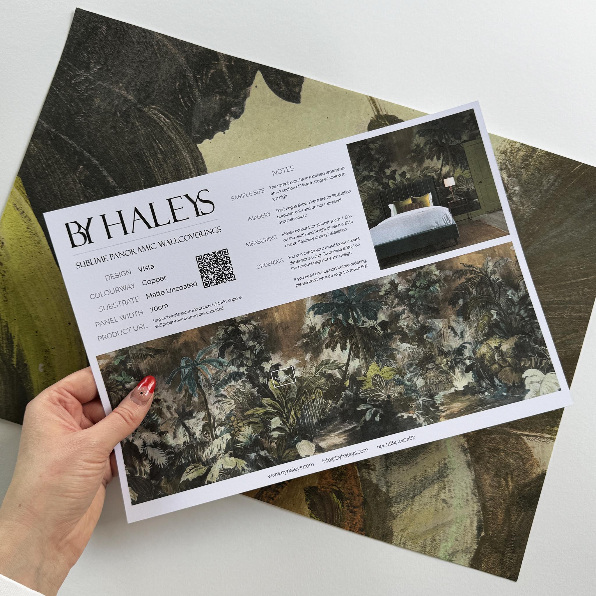 A hand showing information for a matte wallpaper mural sample which features a moody jungle in teal and ochre.
