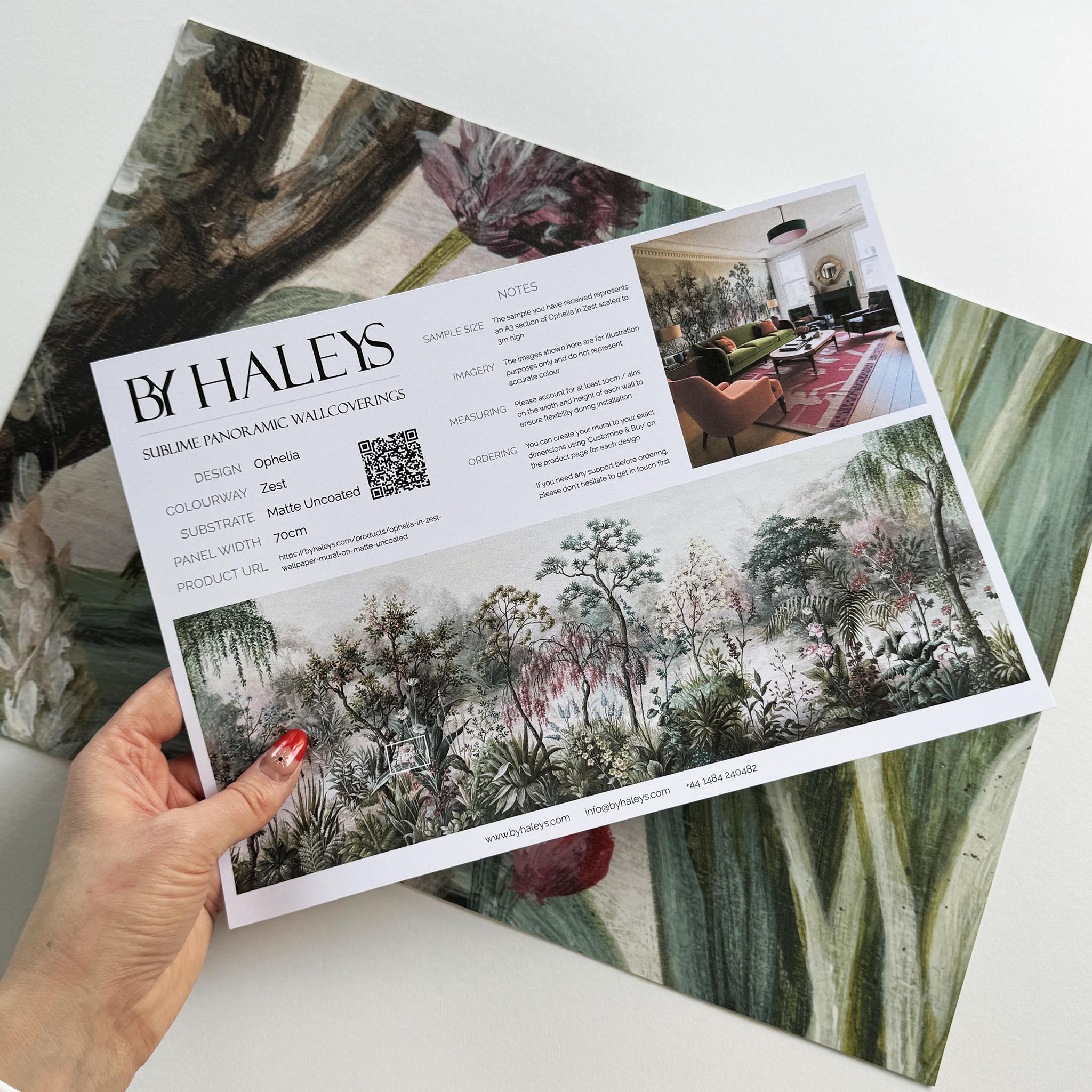 A hand showing information for a matte wallpaper mural sample which features a lakeside garden scene in mint and pink.