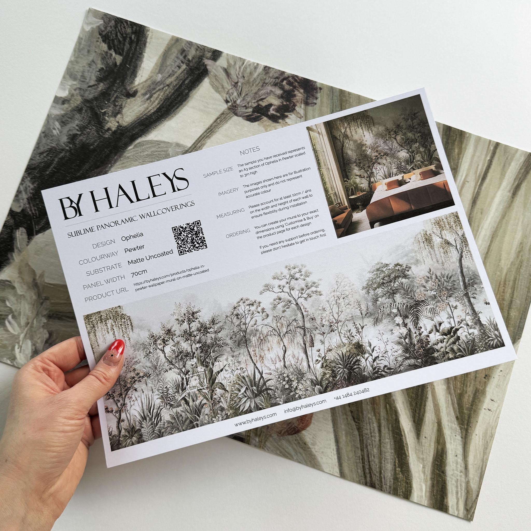 A hand showing information for a matte wallpaper mural sample which features a lakeside scene in grey and peach.