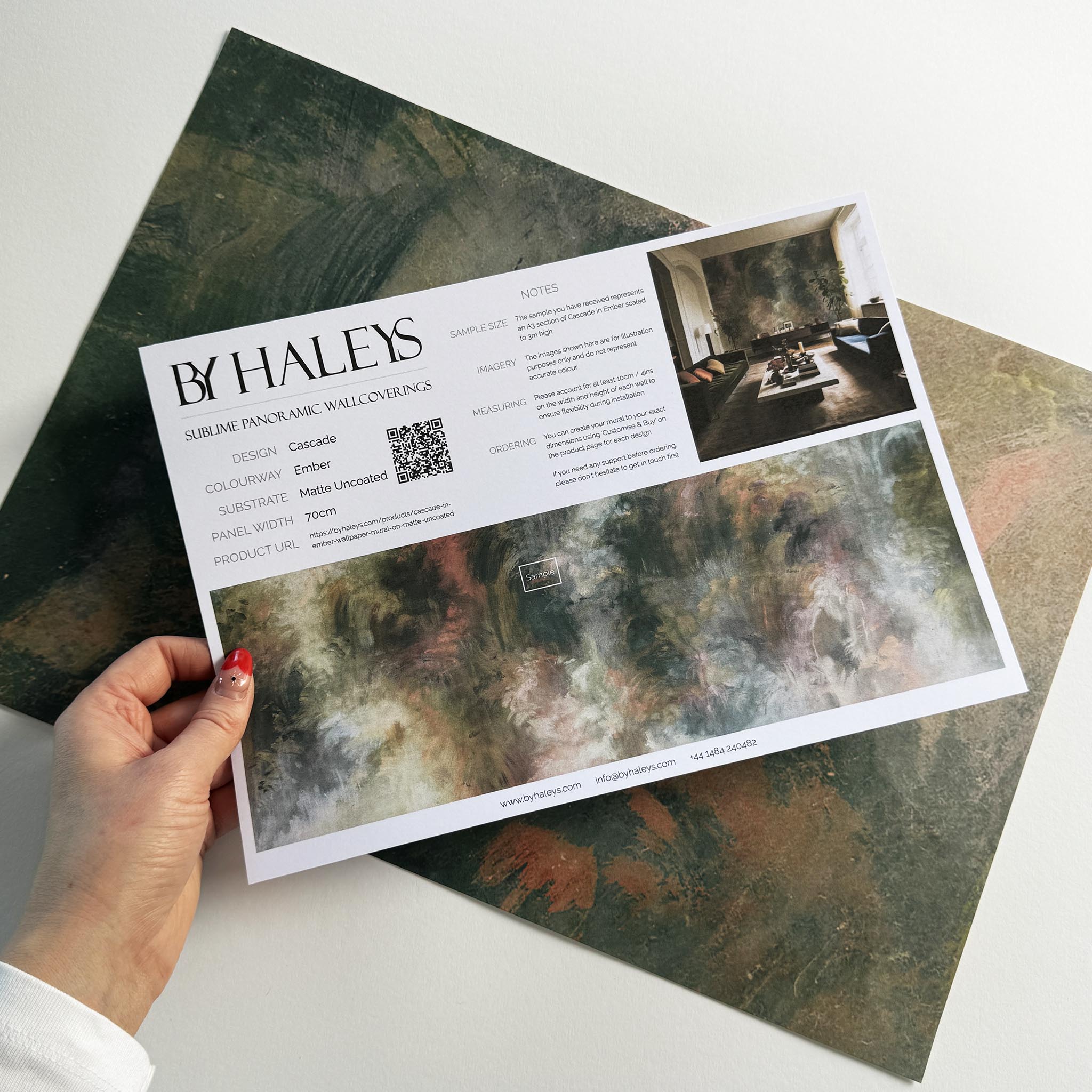 A hand showing information for a matte wallpaper mural sample which features an abstract haze in green and blush.