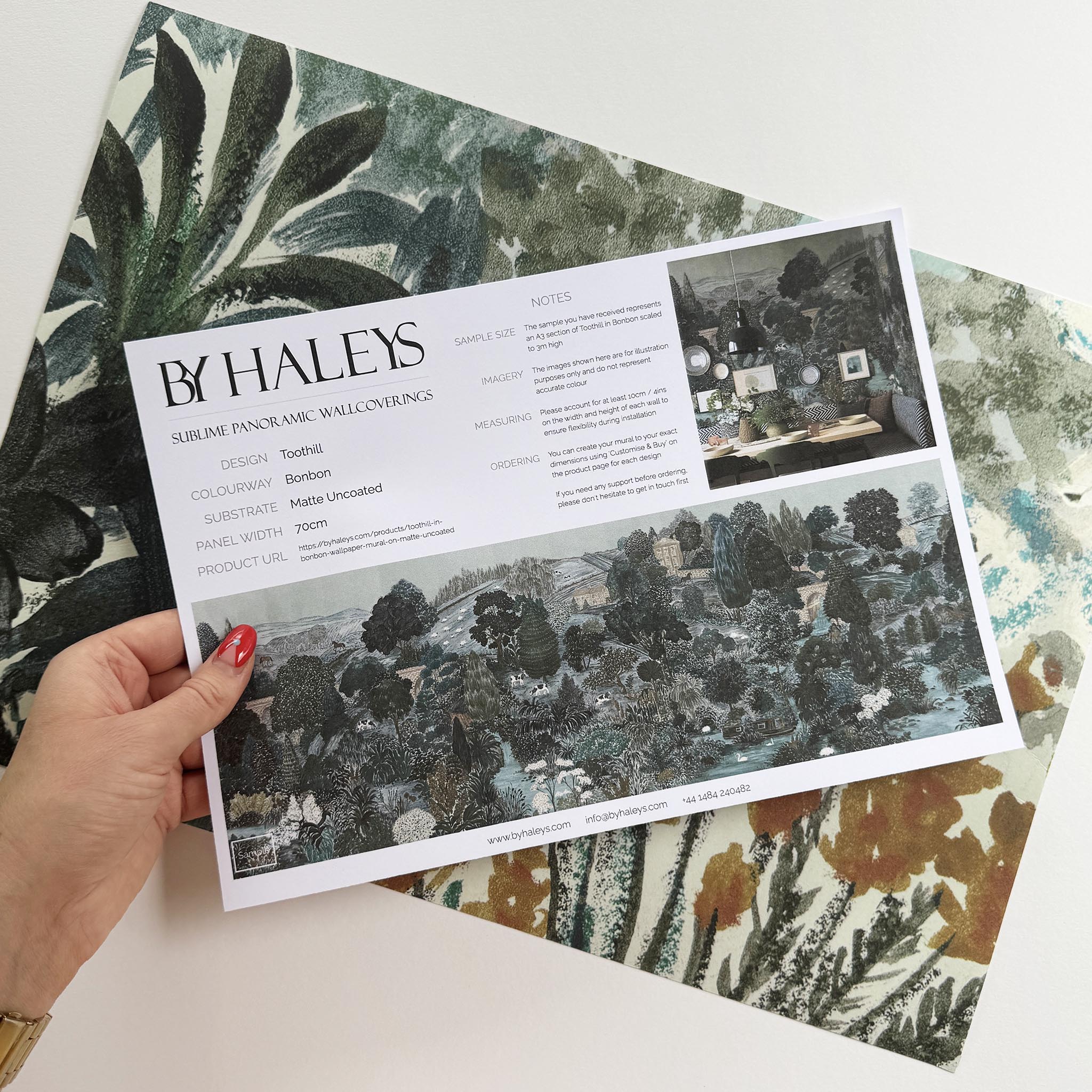 A hand showing information for a matte wallpaper mural sample which features a countryside in teal, sea green and slate 