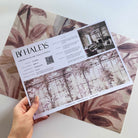 A hand showing information for a pearlescent wallpaper mural sample which features saffron toned mural with climbing foliage and vintage glass.