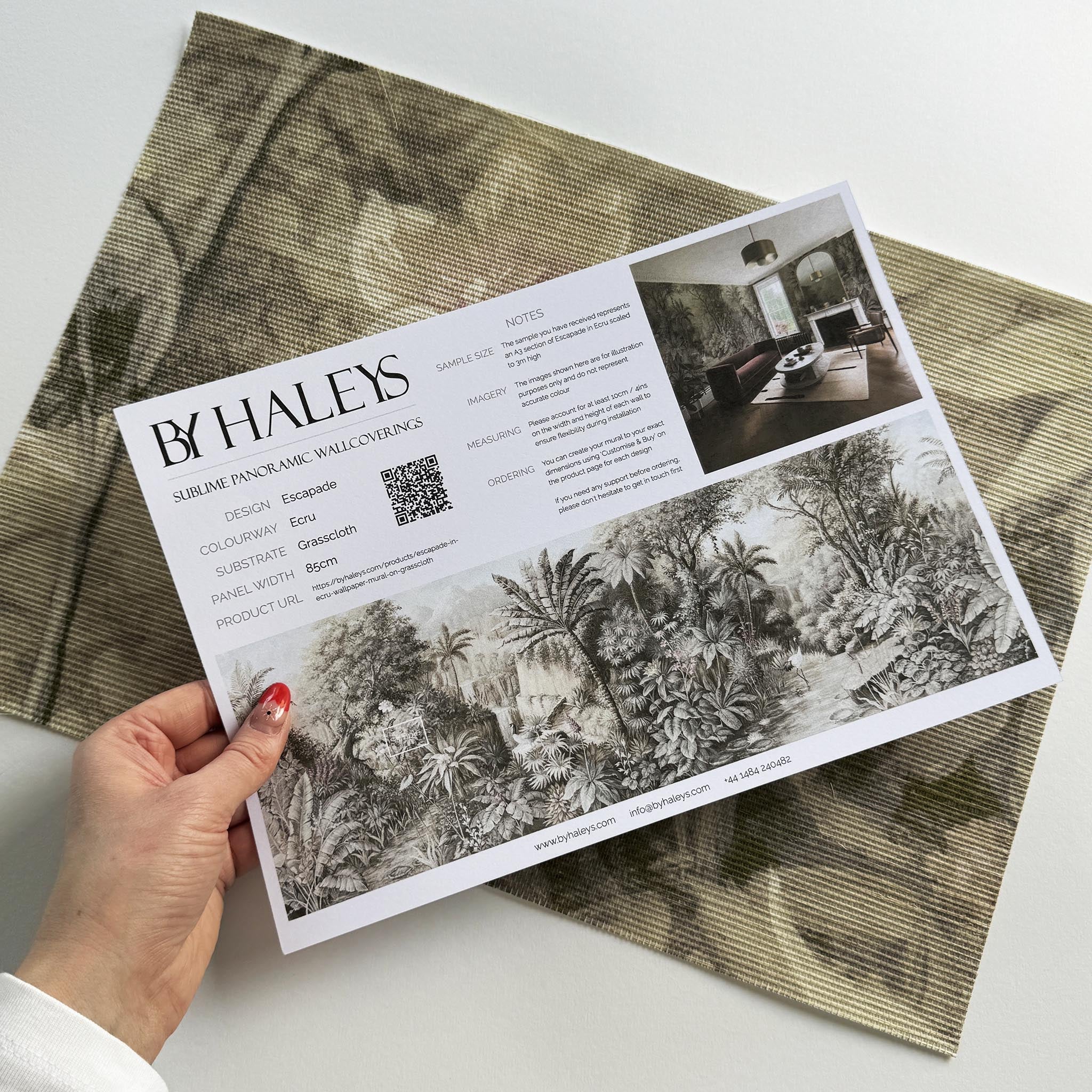 A hand showing information for a grasscloth wallpaper mural sample which features a jungle vista in sepia and blush.