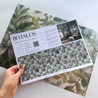 A hand showing information for a pearlescent wallpaper mural sample which features calm mural in chamomile, sage, olive and cream tones.