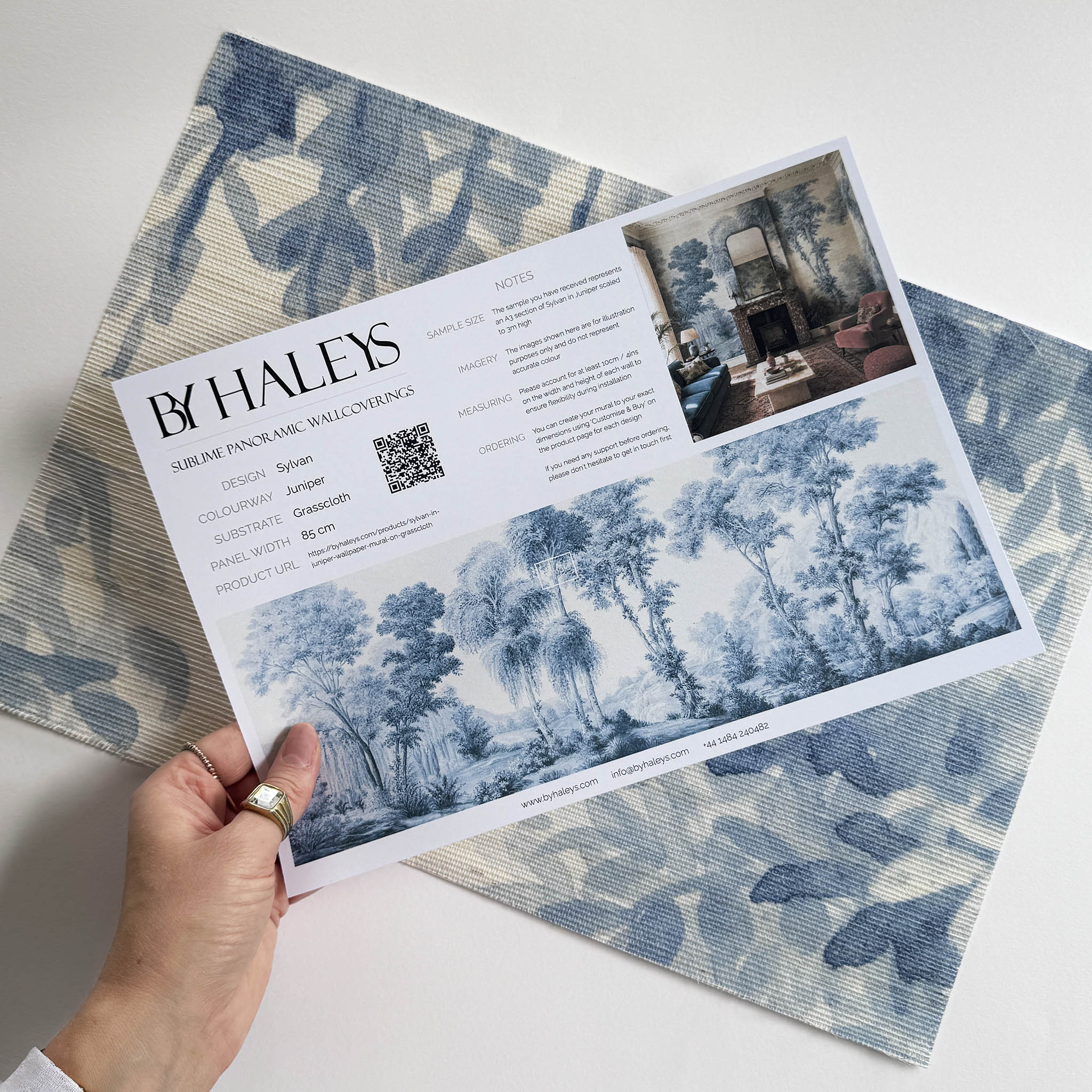A hand showing information for a grasscloth wallpaper mural sample which features soft blue woodland
