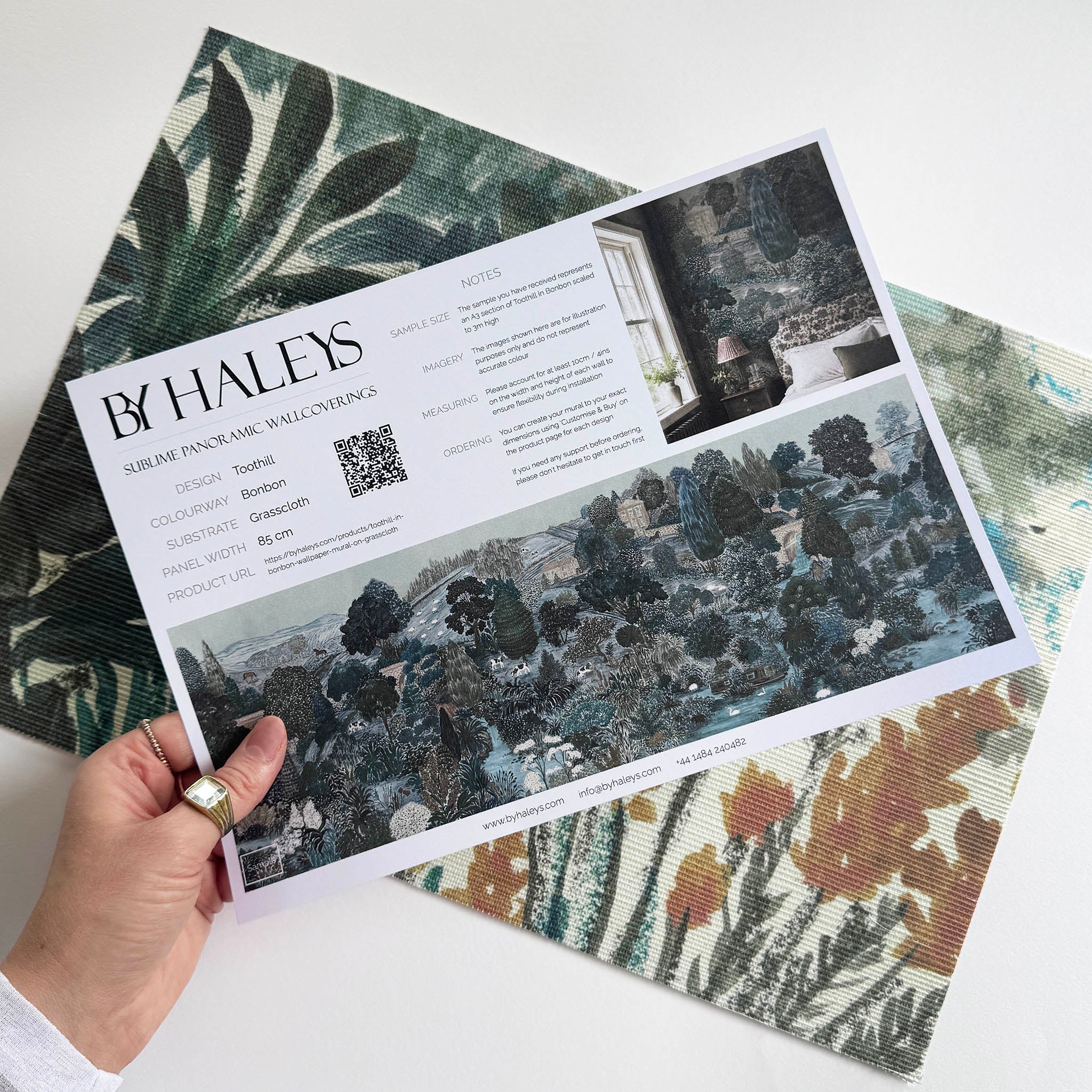 A hand showing information for a grasscloth wallpaper mural sample which features a countryside in teal, sea green and slate 
