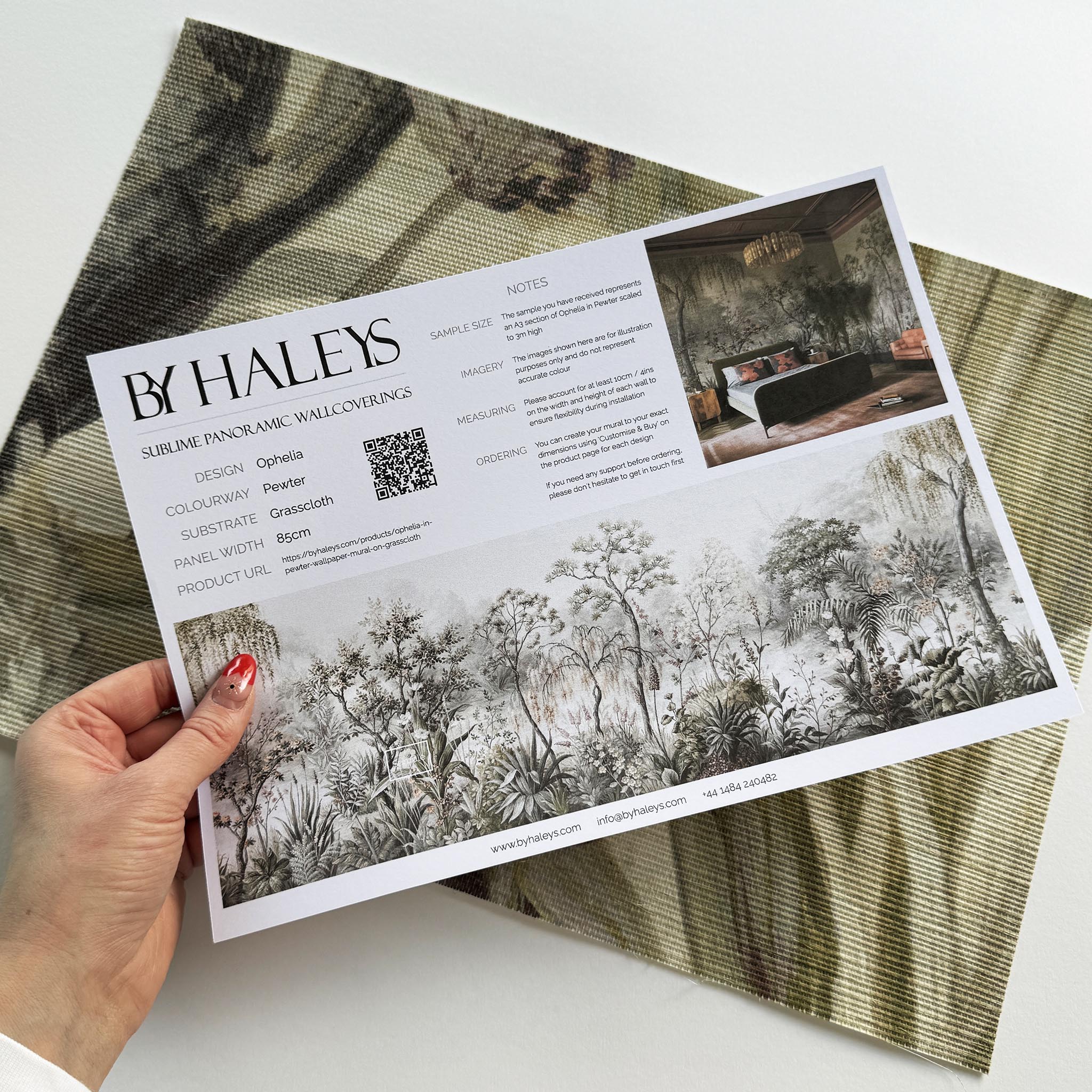 A hand showing information for a grasscloth wallpaper mural sample which features a lakeside scene in grey and peach.