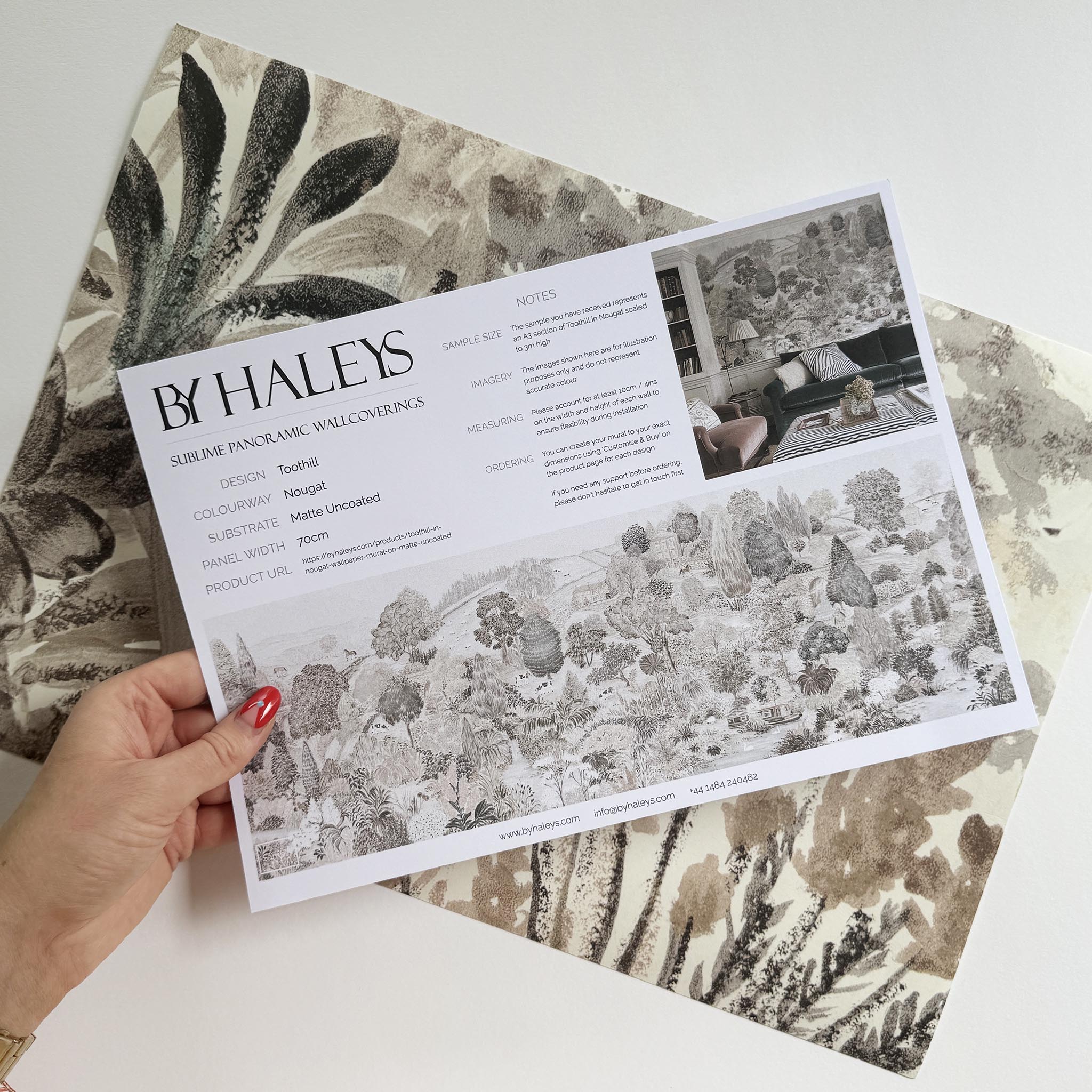 A hand showing information for a matte wallpaper mural sample which features a countryside in almond, dusty rose, and mist
