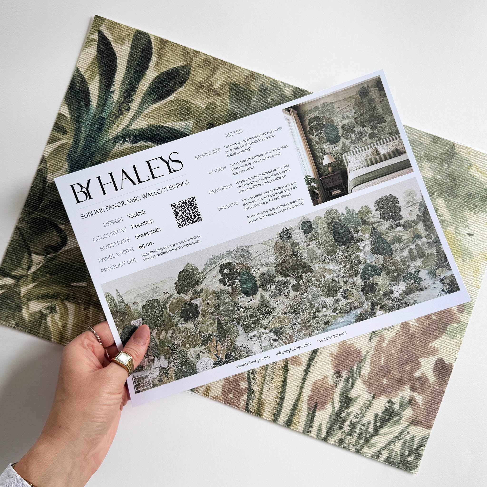 A hand showing information for a grasscloth wallpaper mural sample which features a soft green countryside with cattle and boats
