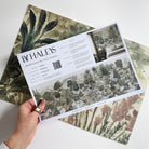 A hand showing information for a grasscloth wallpaper mural sample which features a soft green countryside with cattle and boats