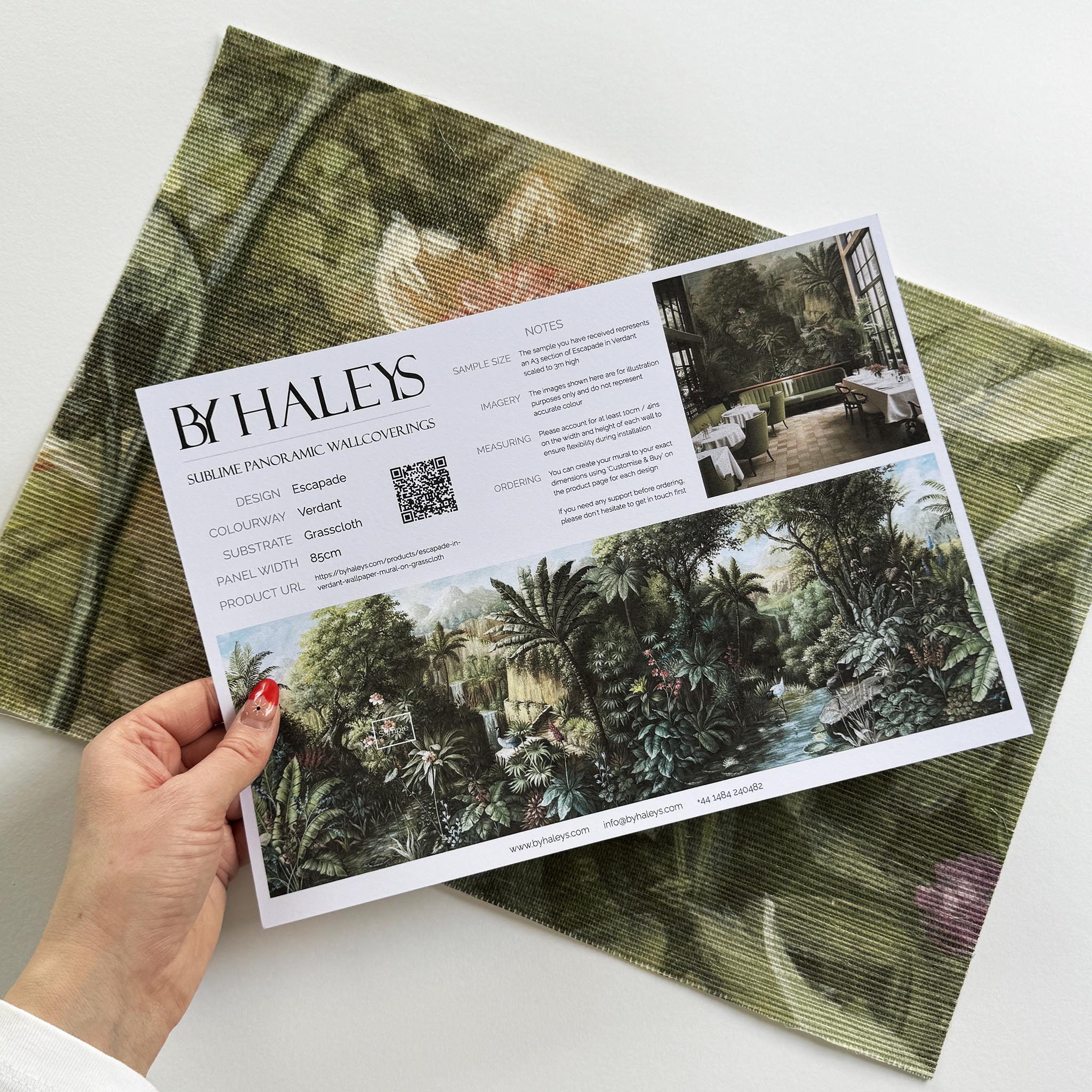 A hand showing information for a grasscloth wallpaper mural sample which features a tropical scene in green and pink.